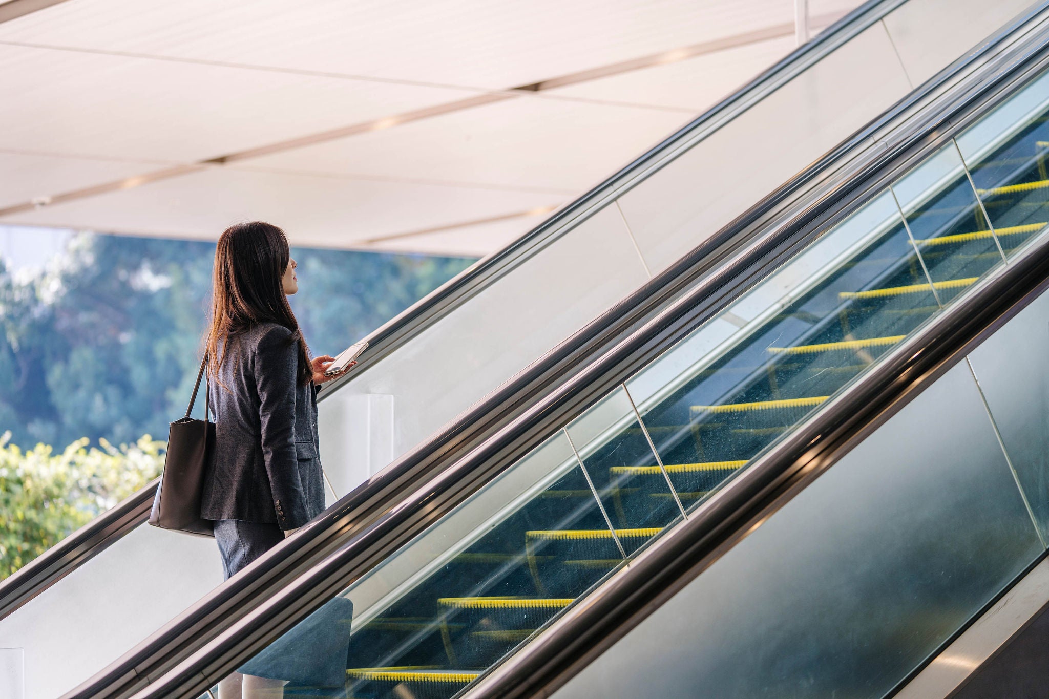 Japanese businesswoman ascending an escalator in a sleek blazer and handbag, symbolizing career progress, urban commuting and confident professional ambition in a modern city interior