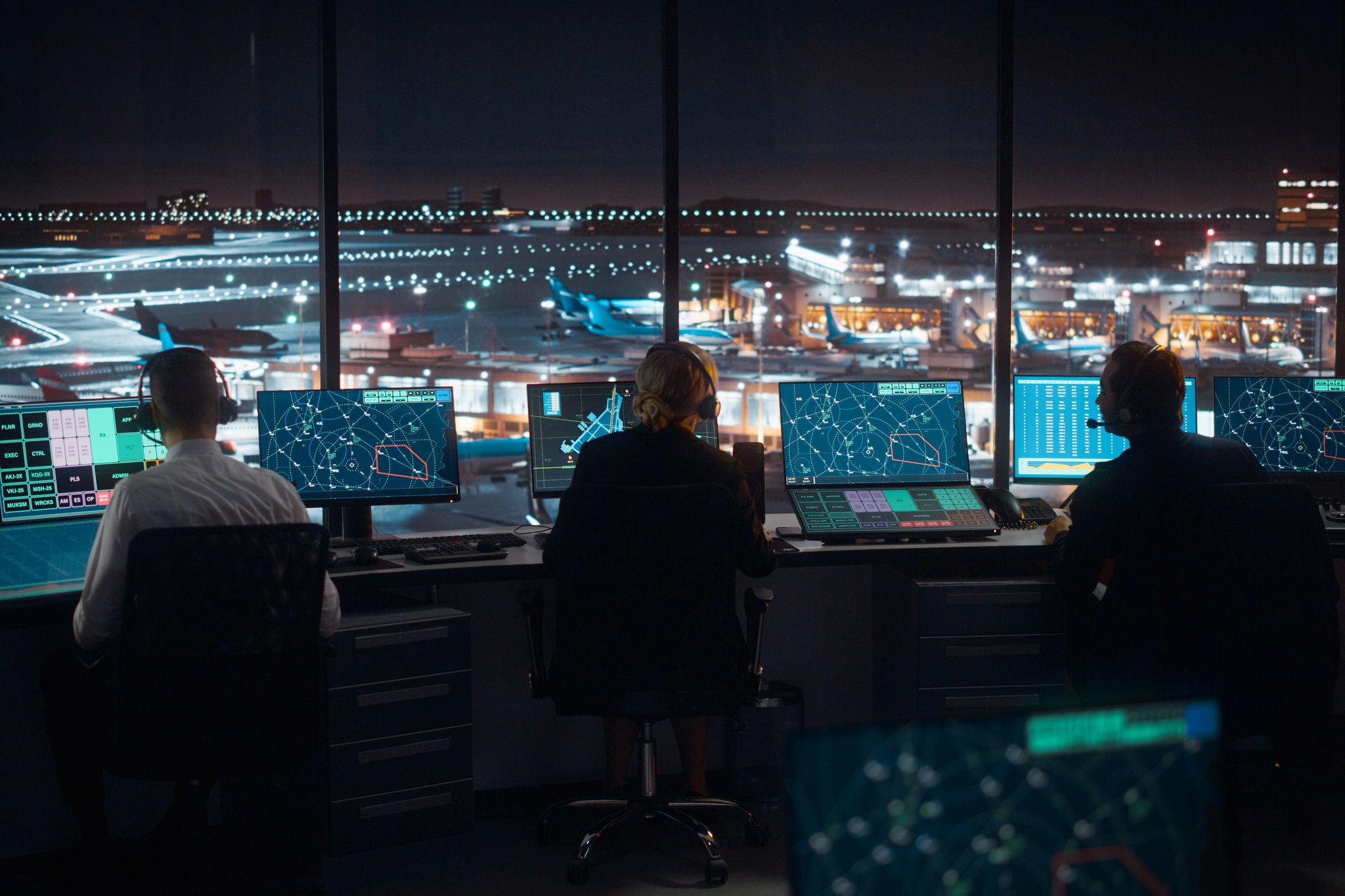 People sit at control desks using multiple screens in an airport tower at night