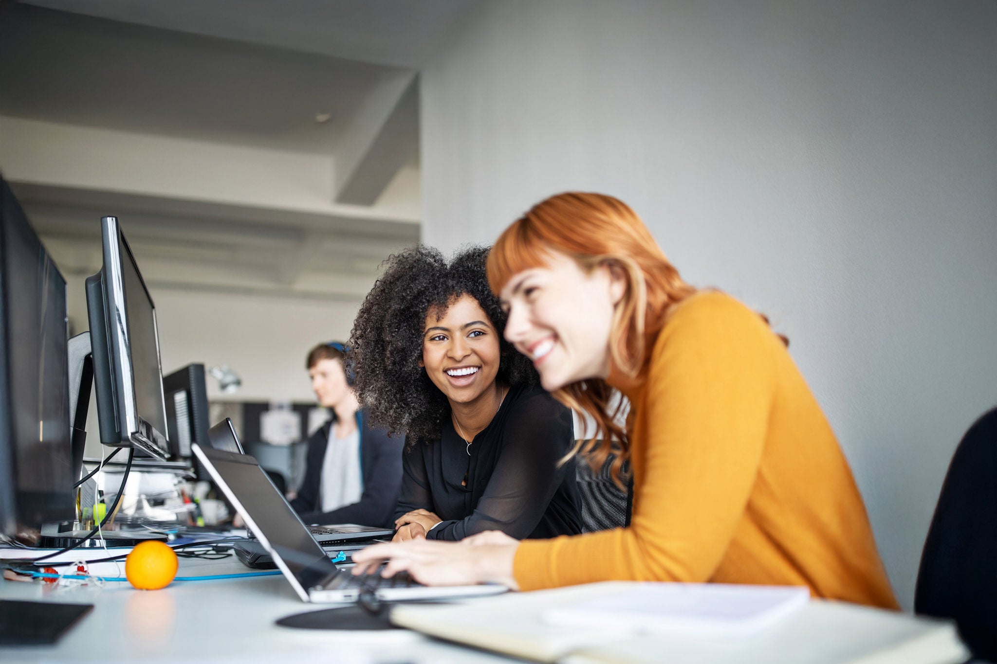 Two young women working together on laptop with male colleague in background. Two female colleagues in office working together.