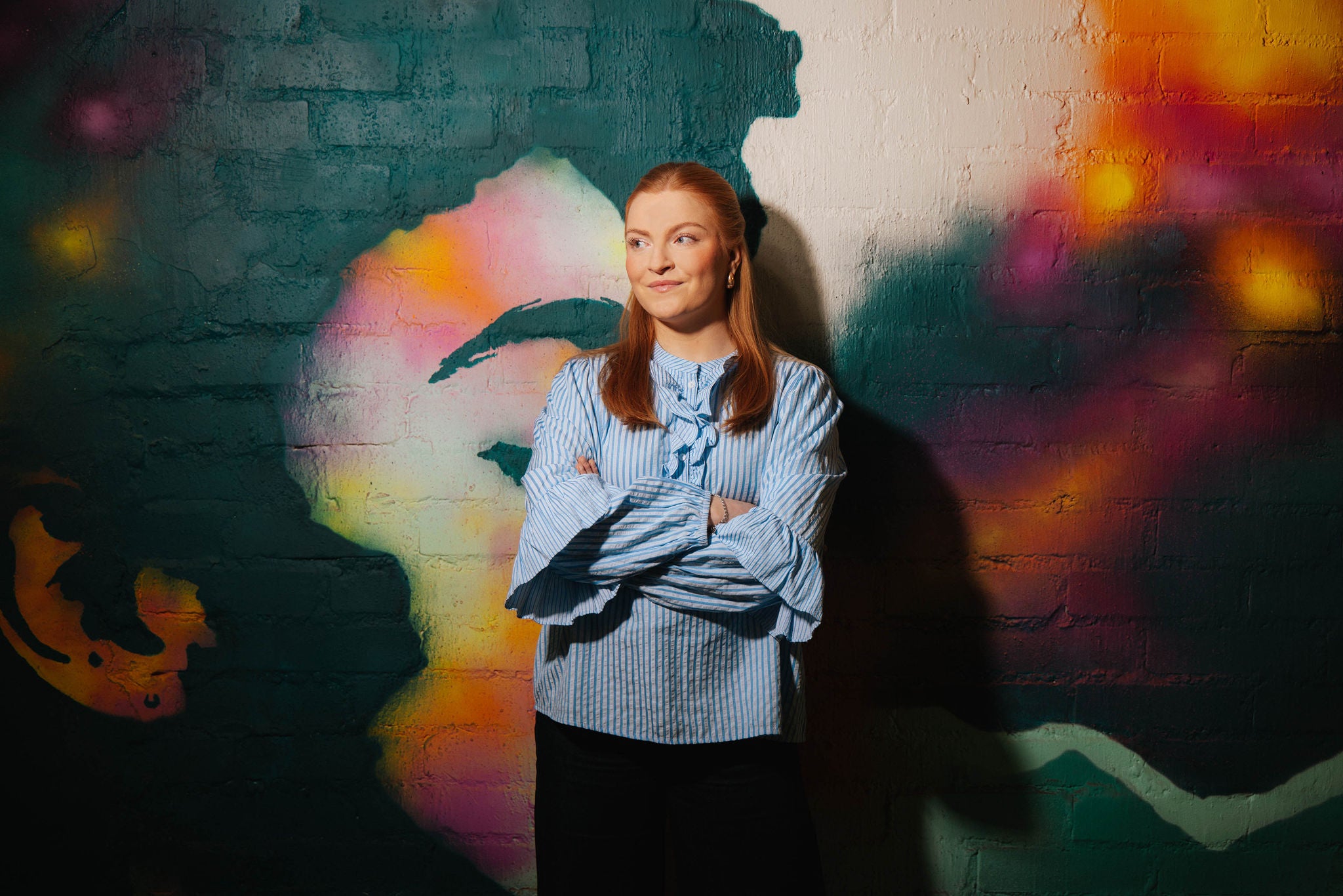 A woman standing with folded arms in front of a colorful mural looking to the left with confidence.