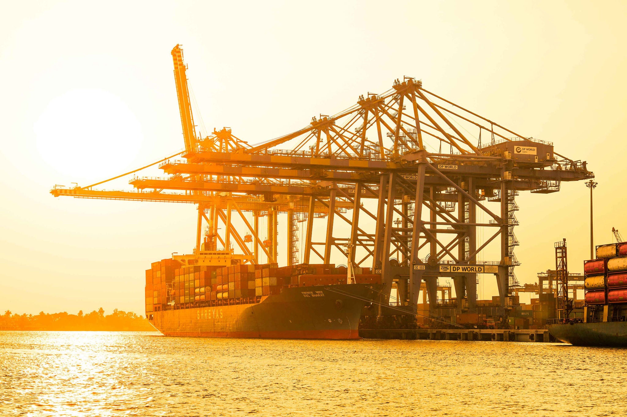 View of a massive cargo ship dwarfed by towering cranes under a golden sky, docked at the bustling port in Kochi, Kerala, India.