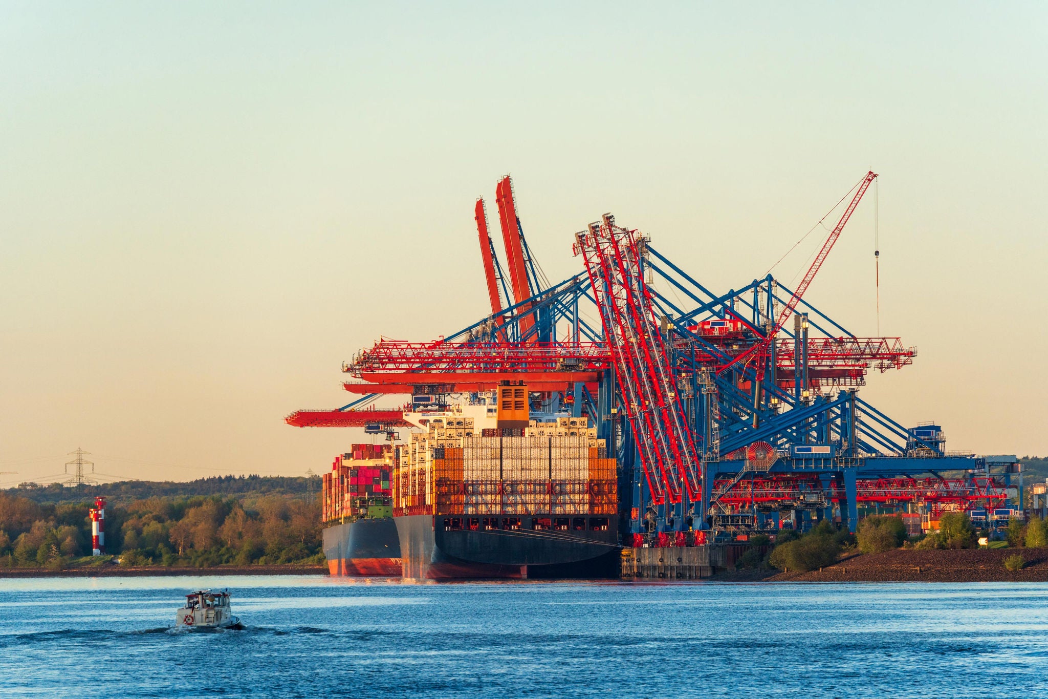 Large container ships docked at a cargo terminal with towering cranes, viewed across a calm waterway.