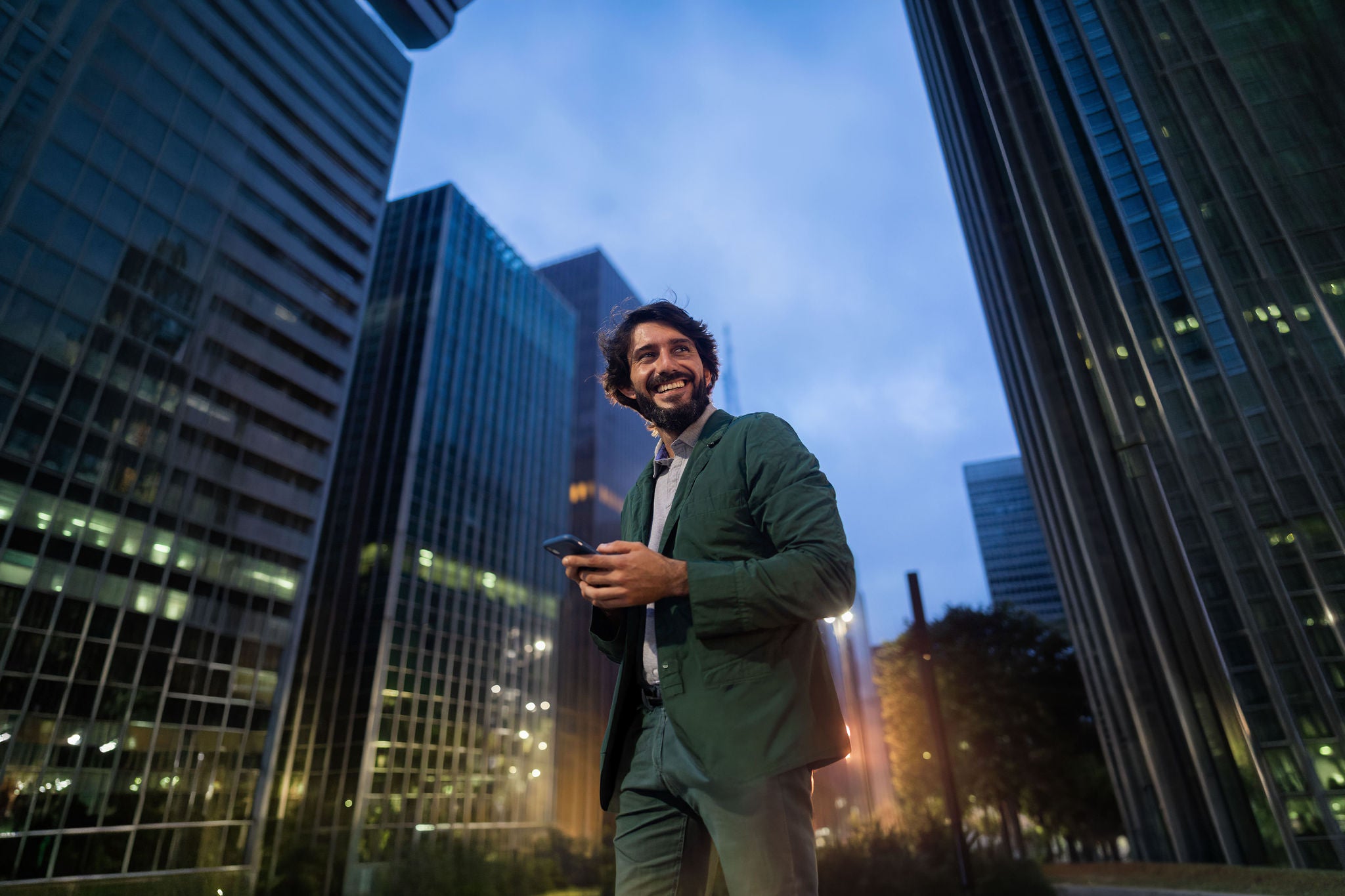 View of young man using a smartphone at night time with city view landscape in the background. Mobile phone, technology, urban concept.