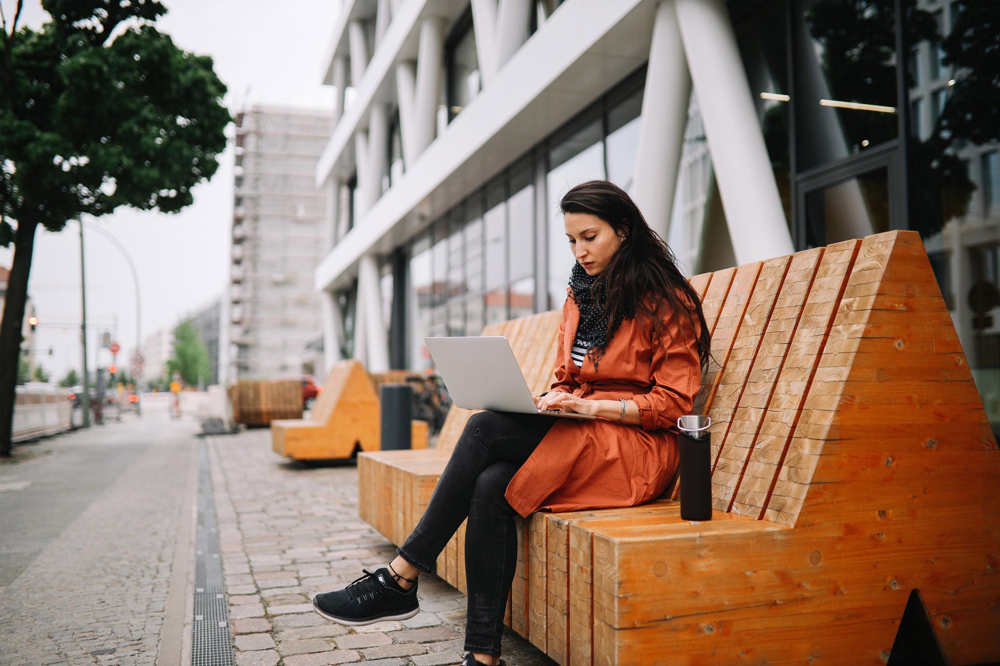 Young commuter woman working on the go in Berlin. She is sitting on the bench while finishing work on the laptop, having a refreshment from her reusable water bottle.