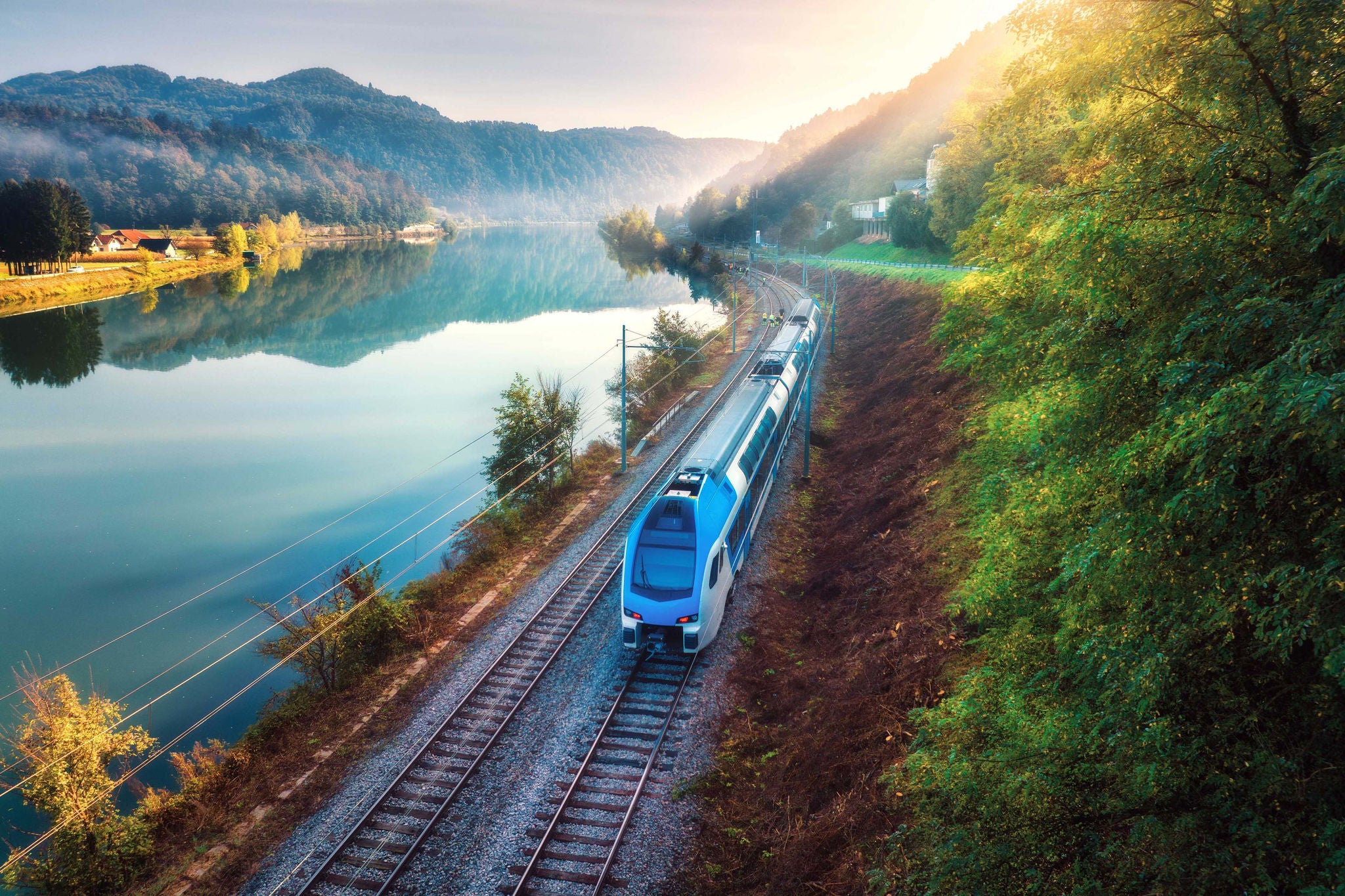 Ein moderner Zug fährt entlang eines ruhigen Flusses, umgeben von Bergen und warmem Morgenlicht.