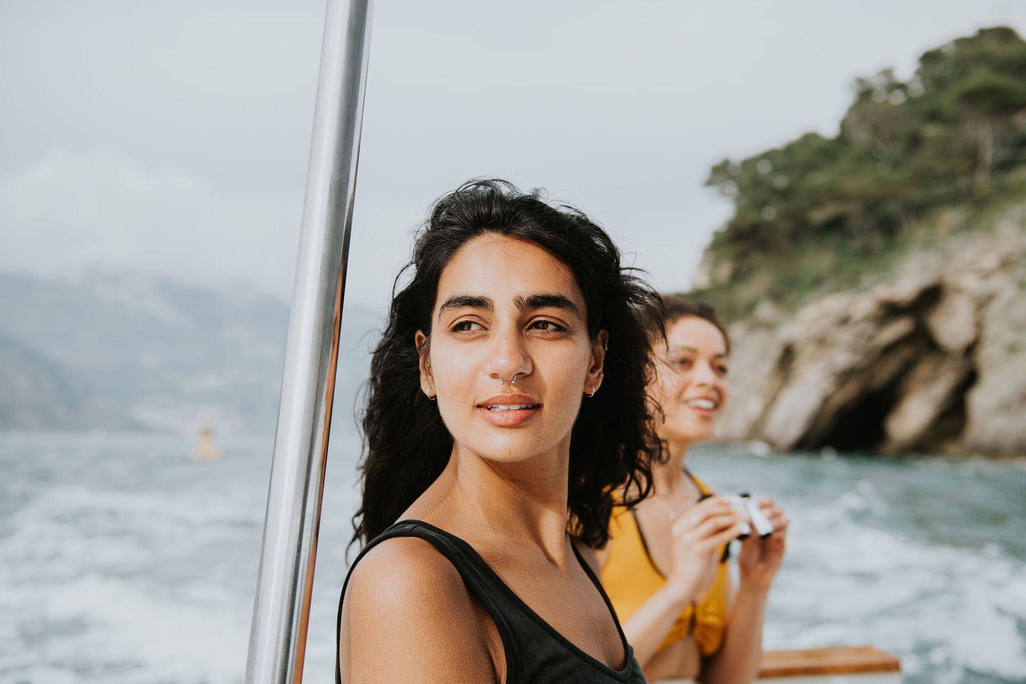 Close-up portrait of a young Indian woman with a nose piercing, on a boat. The sun highlights her skin. She gazes out to sea. Another female passenger is visible behind her.