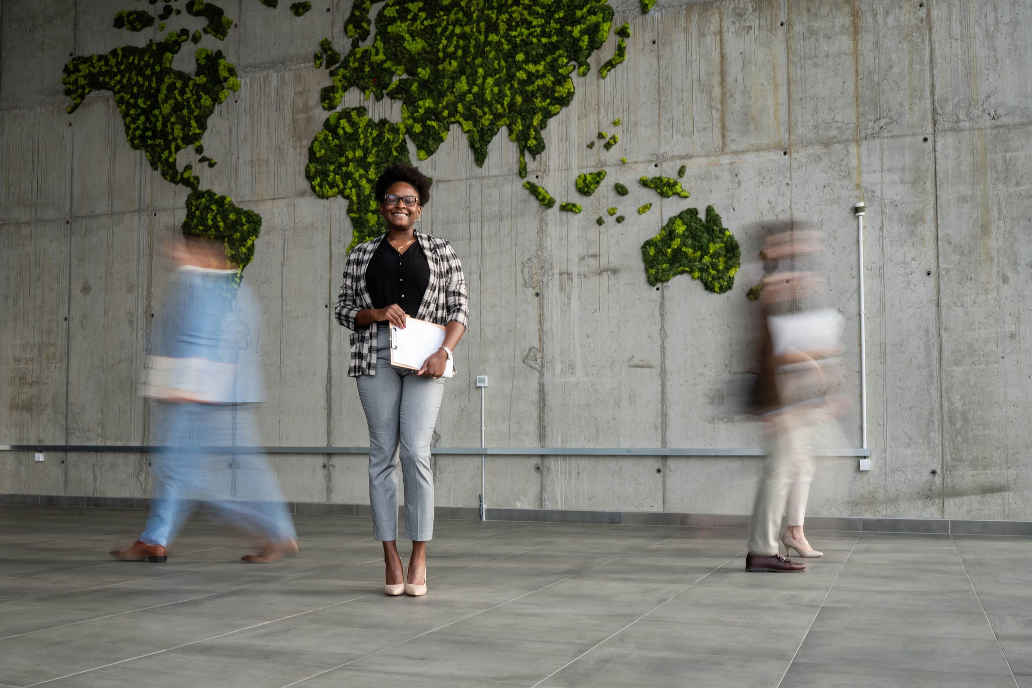 Cheerful businesswoman with tablet standing confidently in a modern office with a green moss world map on a concrete wall