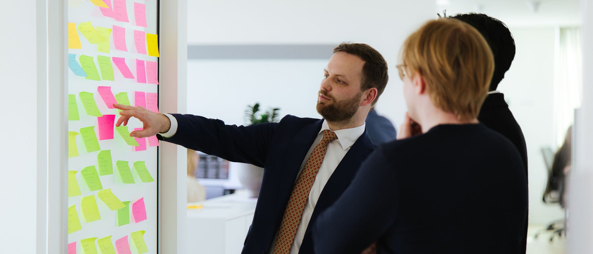 Professionals collaborate around a wall of colorful sticky notes, discussing ideas in an office setting.