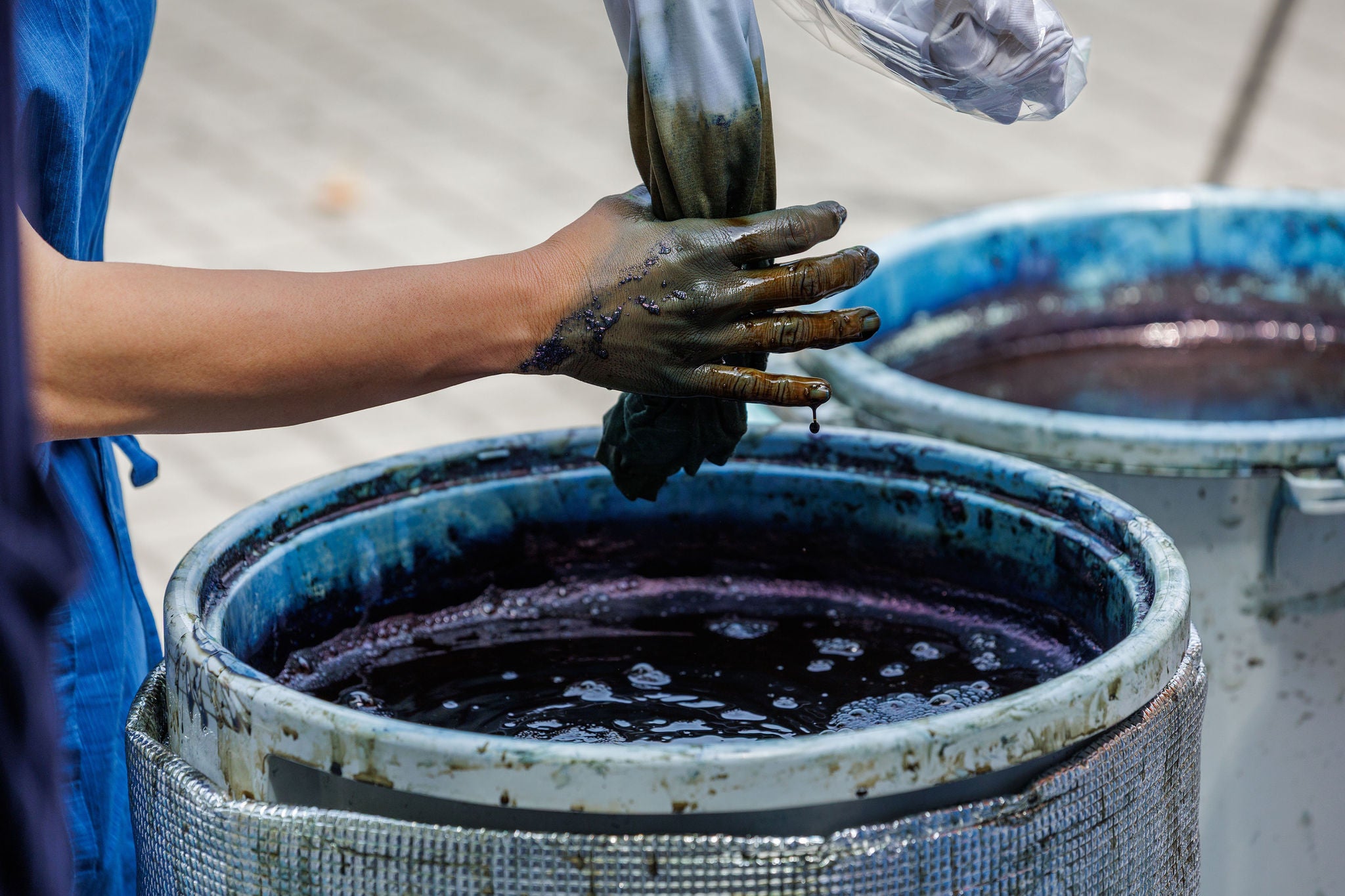 A droplet of blue dye falls from a tanned hand covered in dark indigo liquid from a dipping vat. Scene captures the artistry, texture, and rich hues involved in handmade textile creation.