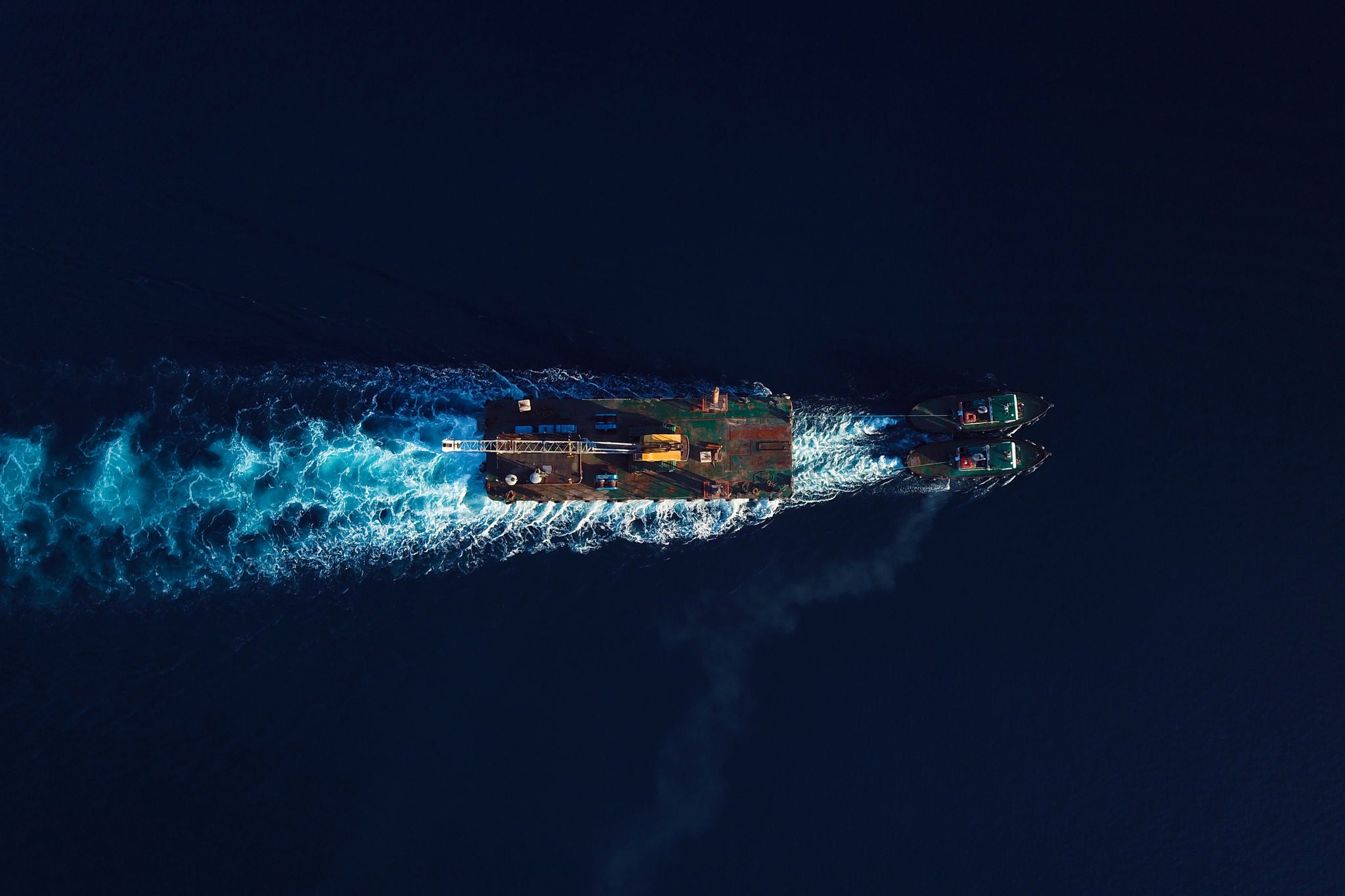 Top-down aerial image of a cargo ship escorted by tugboats on deep blue ocean.