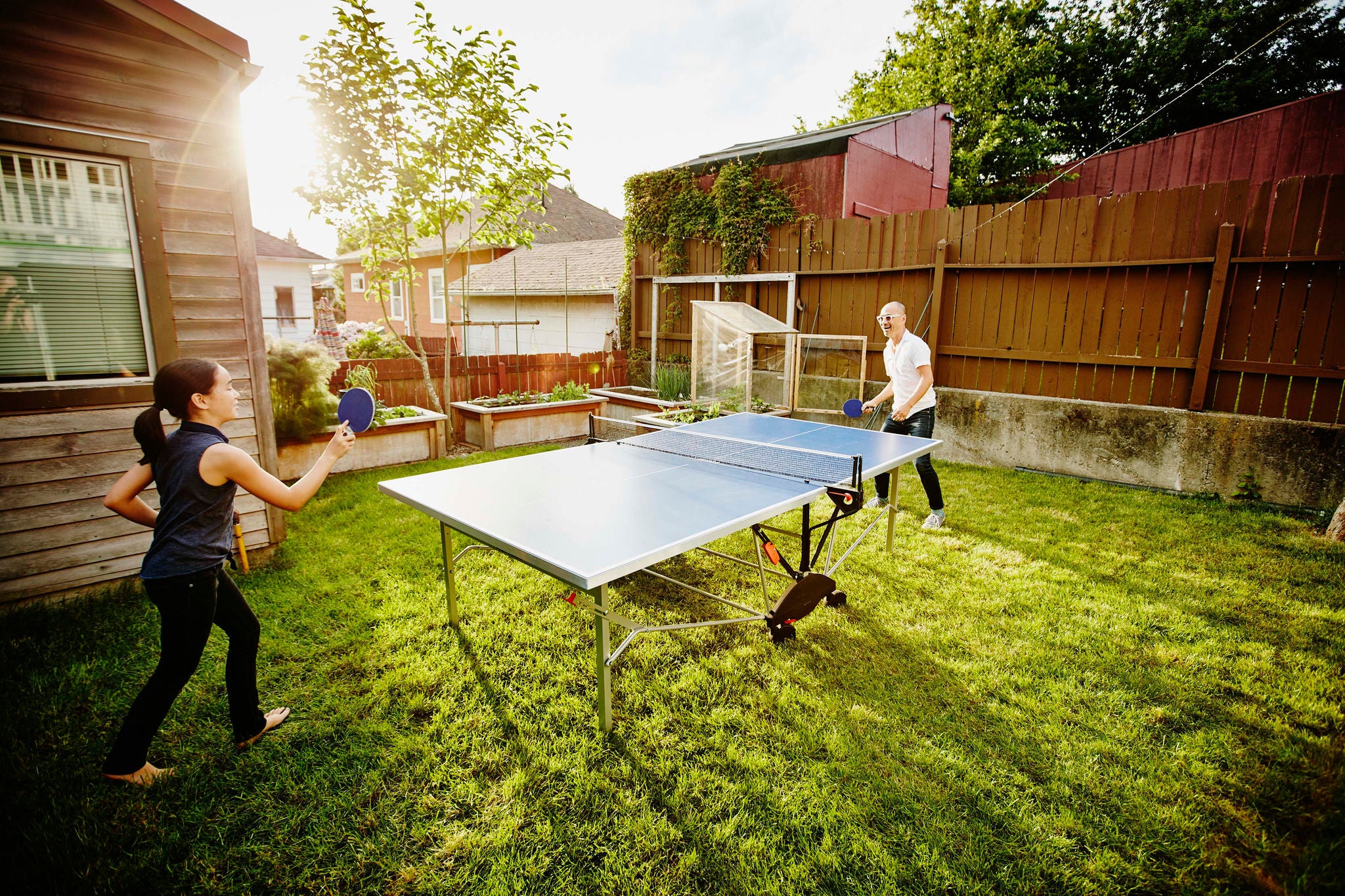 Smiling young daughter and father playing ping pong in backyard of home on summer evening