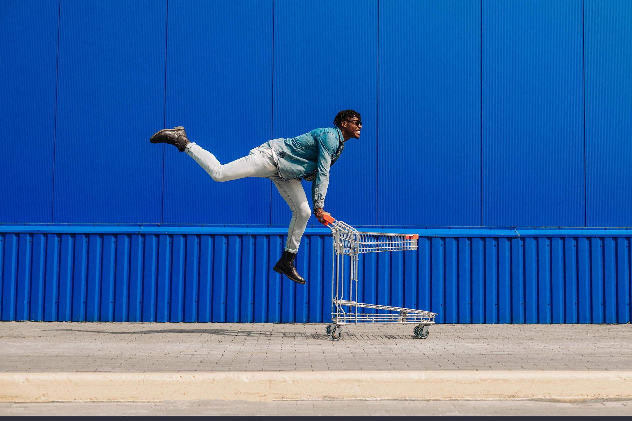 Young happy african american man in casual wear and sunglasses running with empty shopping cart outdoors against blue wall with copy space. Shopping, consumerism concept