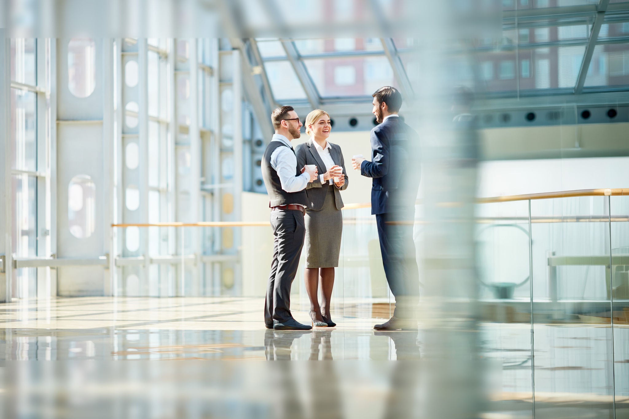 Group of modern business people chatting during coffee break  standing in sunlit glass hall of office building
