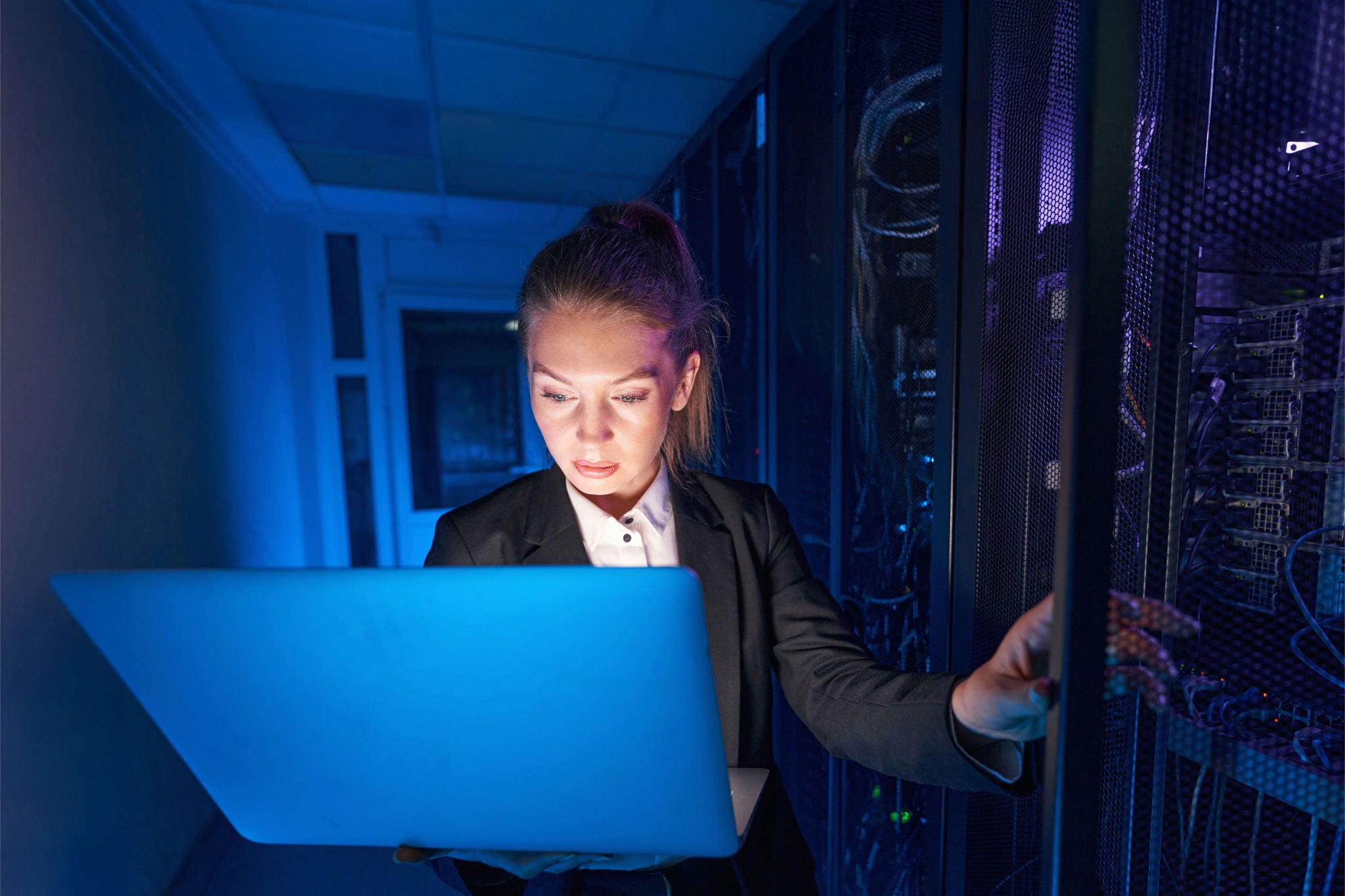 Attractive female IT engineer holding laptop while working in data center with rows of server racks and computers