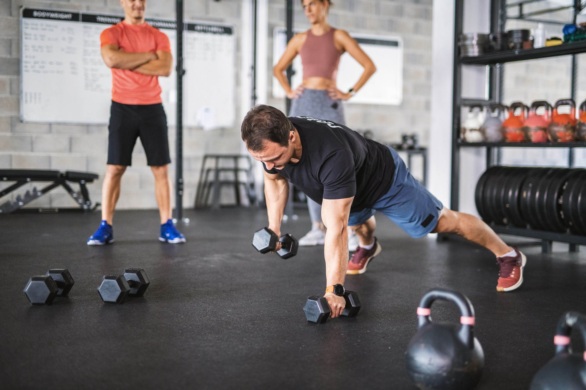 Blick aus der Froschperspektive auf Sportler mittleren Alters in einem Fitnessstudio, die hinter einem Trainer stehen und ihm dabei zusehen, wie er mit gespreizten Beinen Liegestütze mit Hanteln macht.