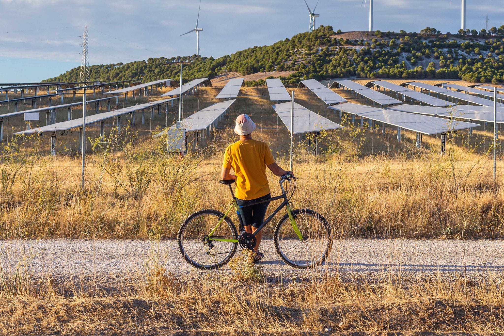 Castilla y Leon, Spain. Man from behind leaning on bicycle looking at wind power towers and solar farm in rural setting at sunset