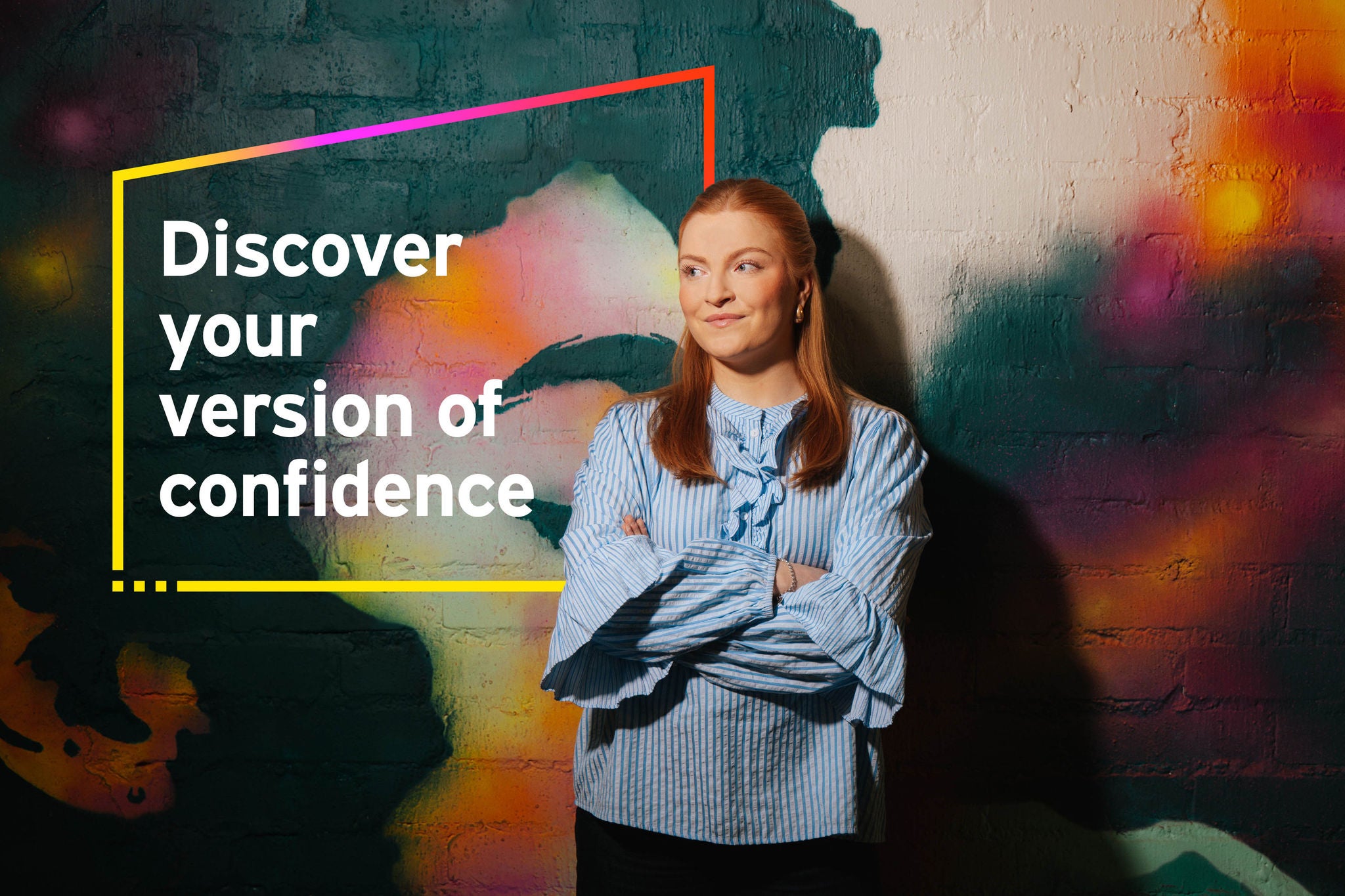 A woman standing with folded arms in front of a colorful mural with the text “Discover your version of confidence.”