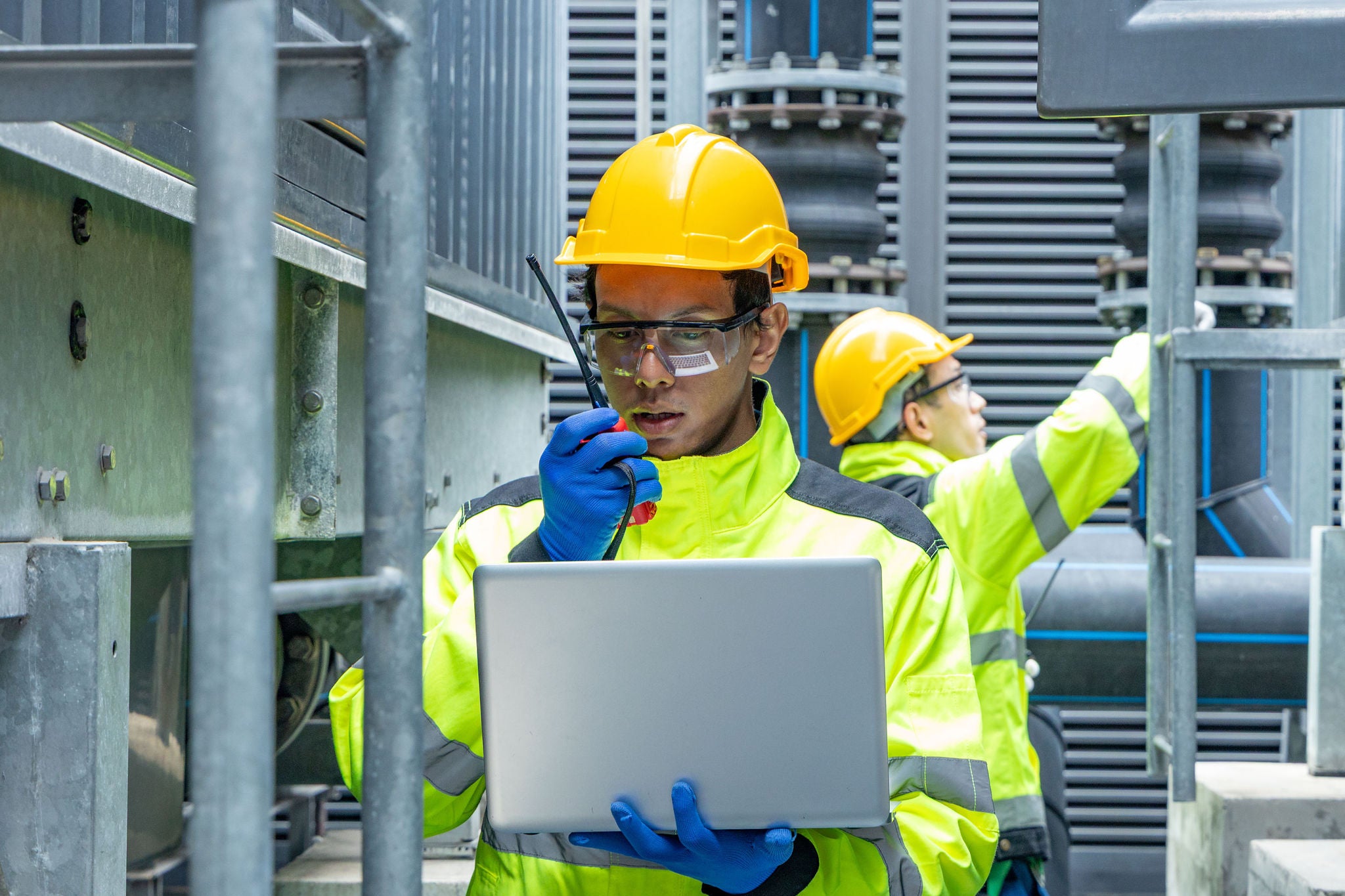 Two engineers in high-visibility gear and helmets examine a control panel on industrial HVAC equipment, using a walkie-talkie to coordinate.