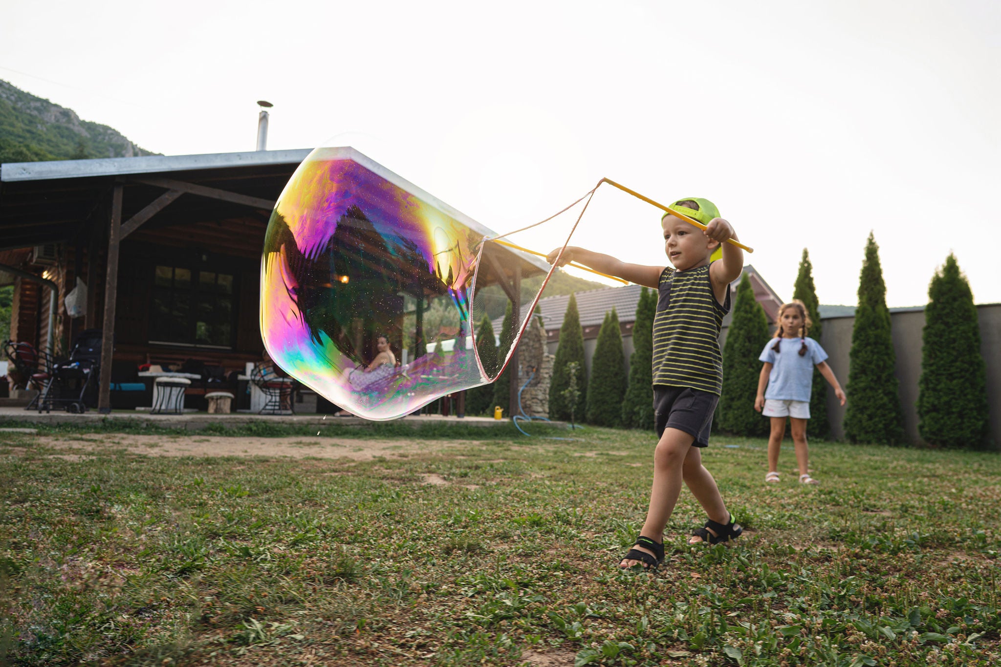 Brother and sister playing together and making a big soap bubble with a bubble wand, during family gathering at the backyard of country house