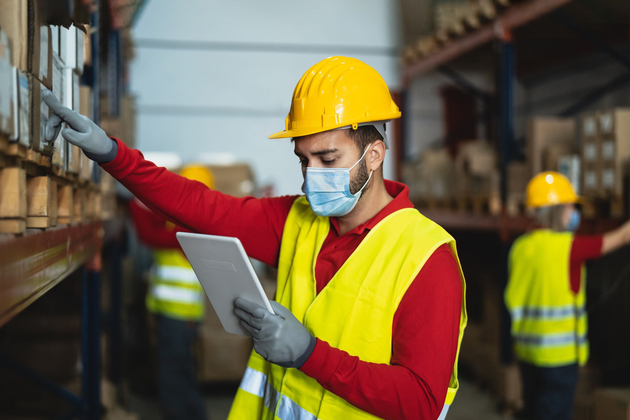 Young man working in warehouse doing inventory using digital tablet and loading delivery boxes plan while wearing face mask during corona virus pandemic - Logistic and industry concept