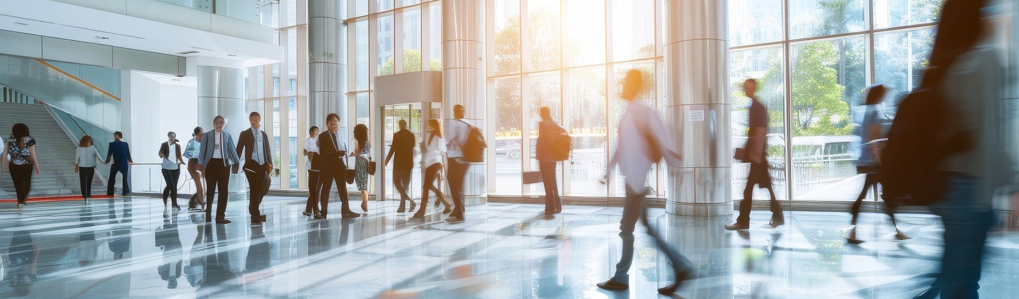 Numerous professionals are walking through a spacious office lobby during a sunny afternoon, showcasing an active environment filled with movement and energy.