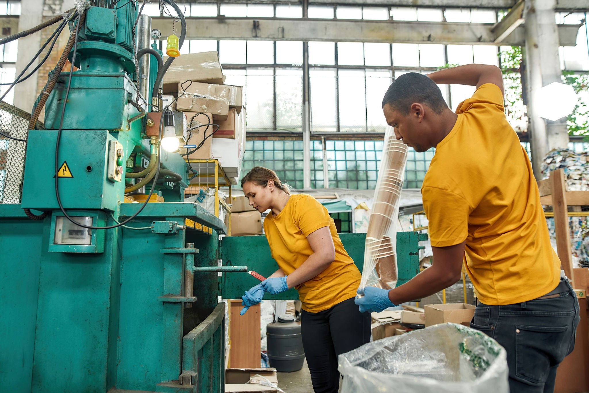 Two workers in a factory handling cardboard and operating machinery.