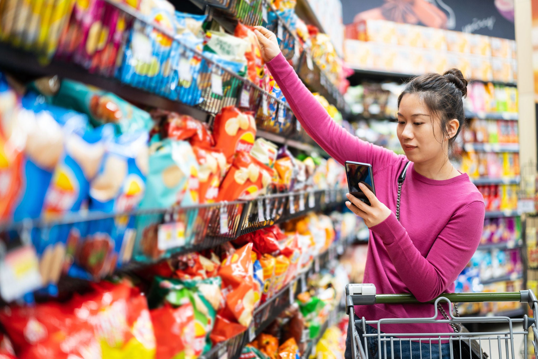 Woman shopping for snacks while using a smartphone in a supermarket