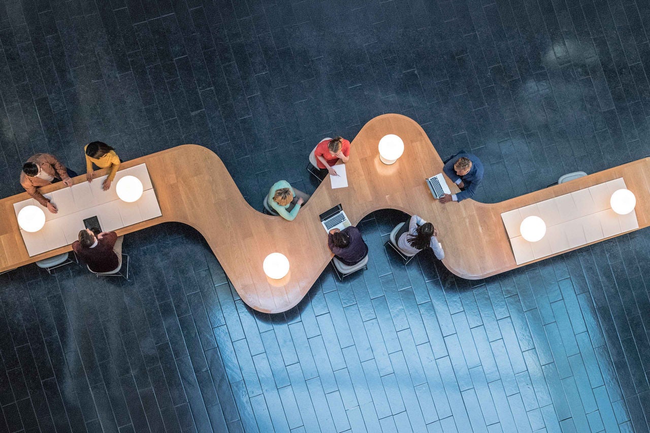 Panoramic overhead view of several business meetings going on in the communal area of a modern office building.