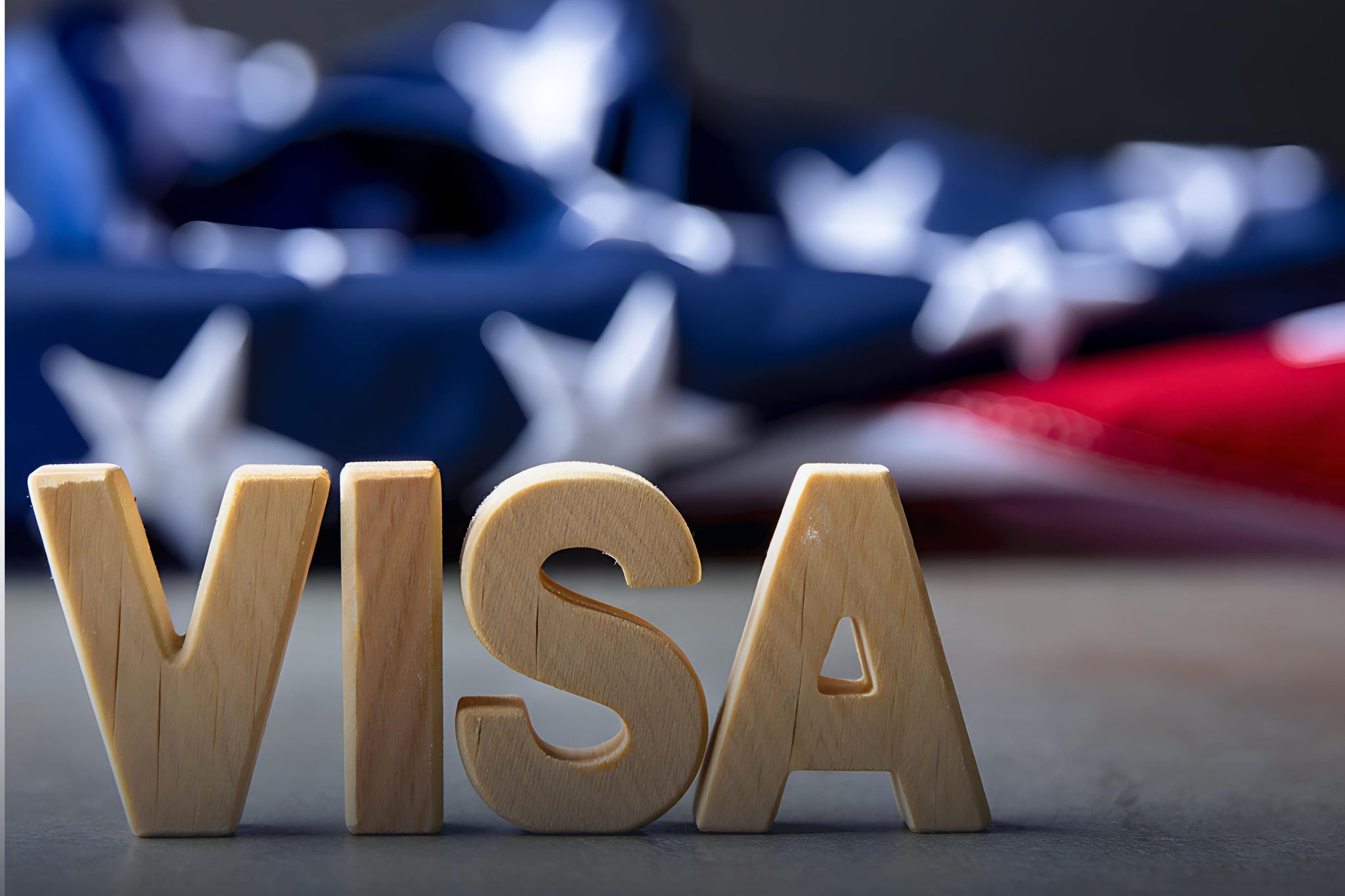 Buchstaben "VISA", die aus Holz gefertigt sind und auf einer Oberfläche liegen. Im Hintergrund ist eine unscharfe Darstellung einer USA-Flagge zu sehen.