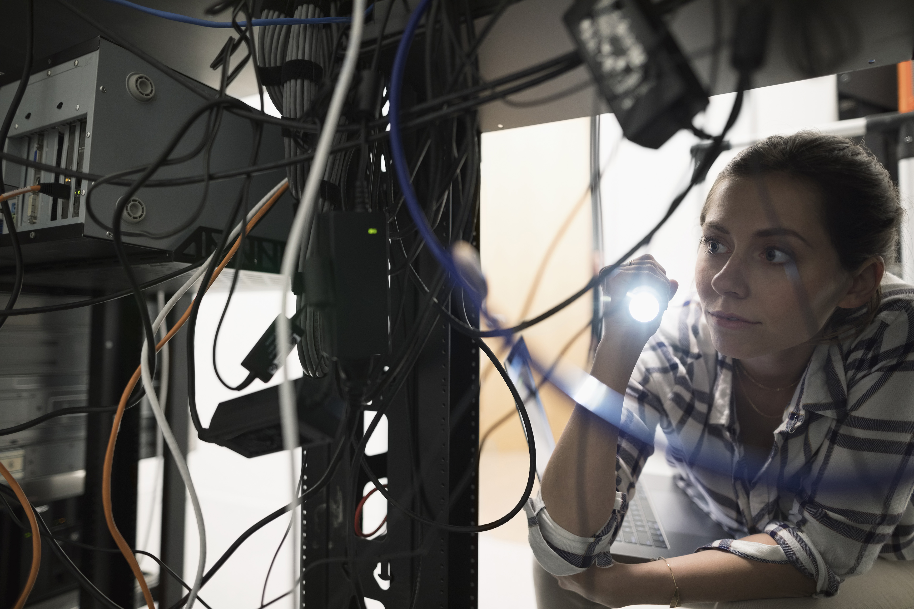 Kvindelig tekniker lyser med lommelygte blandt løse og rodede computerkabler