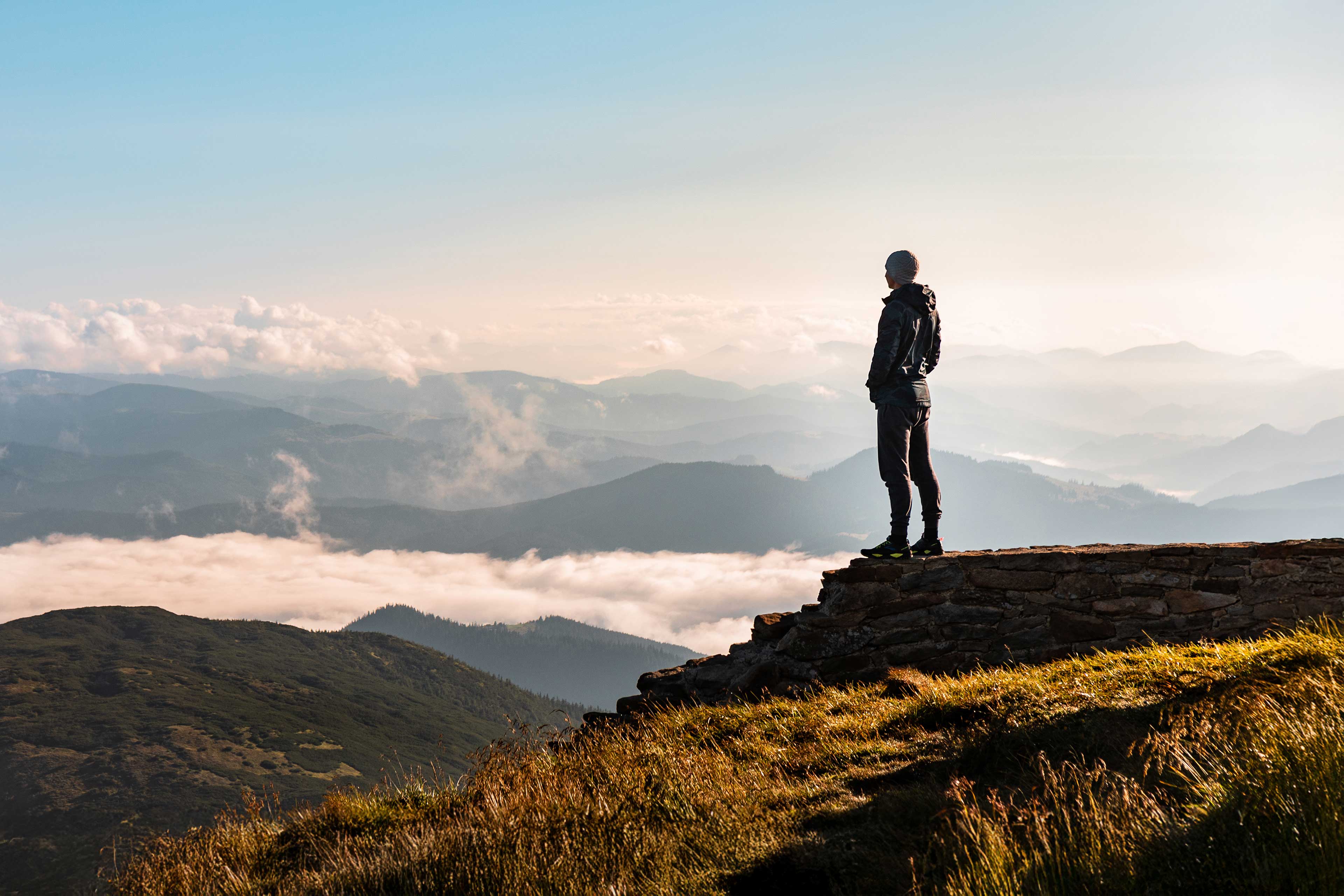 Person steht auf Berggipfel und blickt über ein wolkenverhangenes Tal in die Ferne.