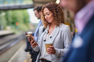 Frau in Business-Kleidung am Bahnhof schaut auf ihr Smartphone und hält einen Kaffeebecher.