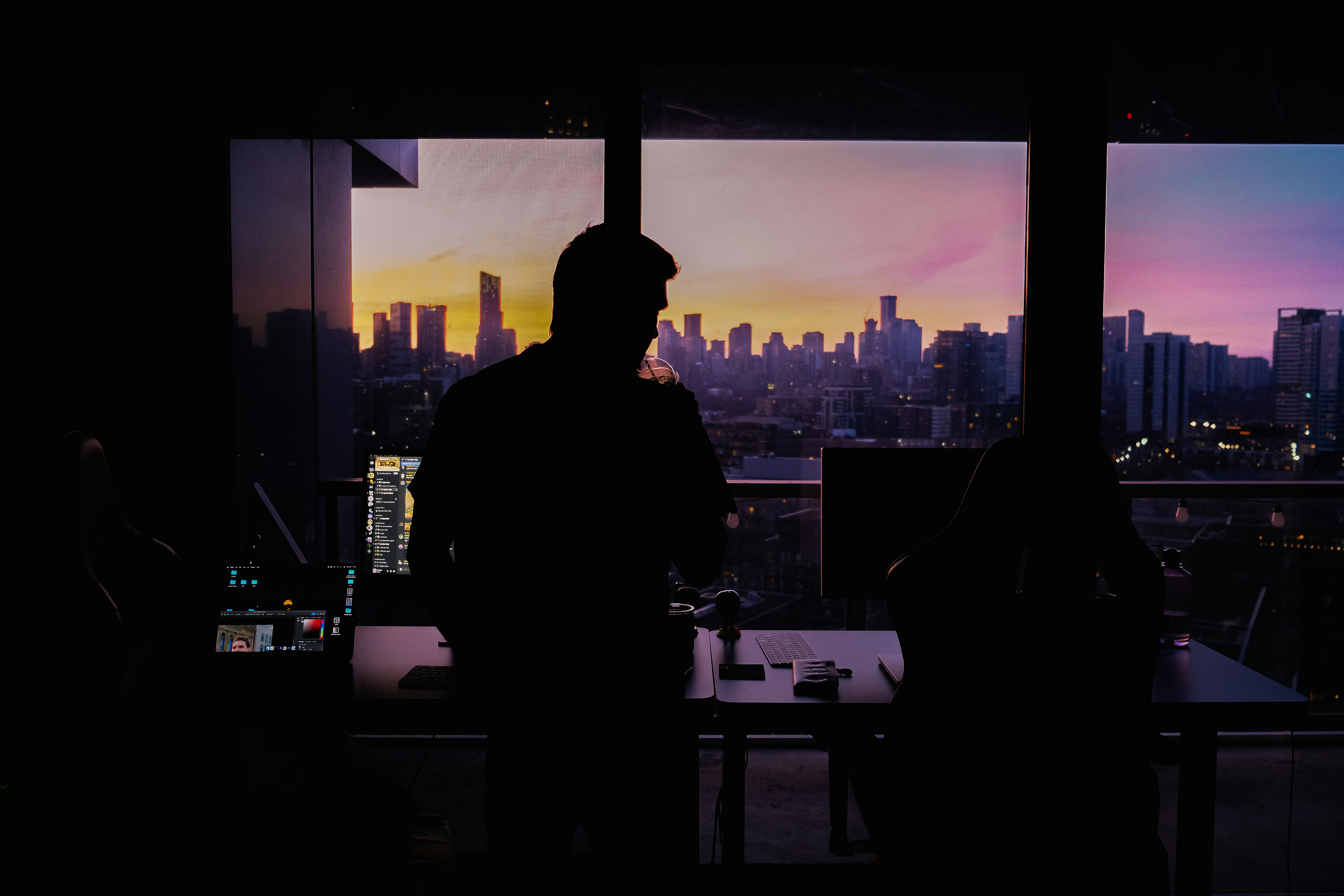 Silhouette of a person in a modern office holding a cup, overlooking a city skyline at sunset.