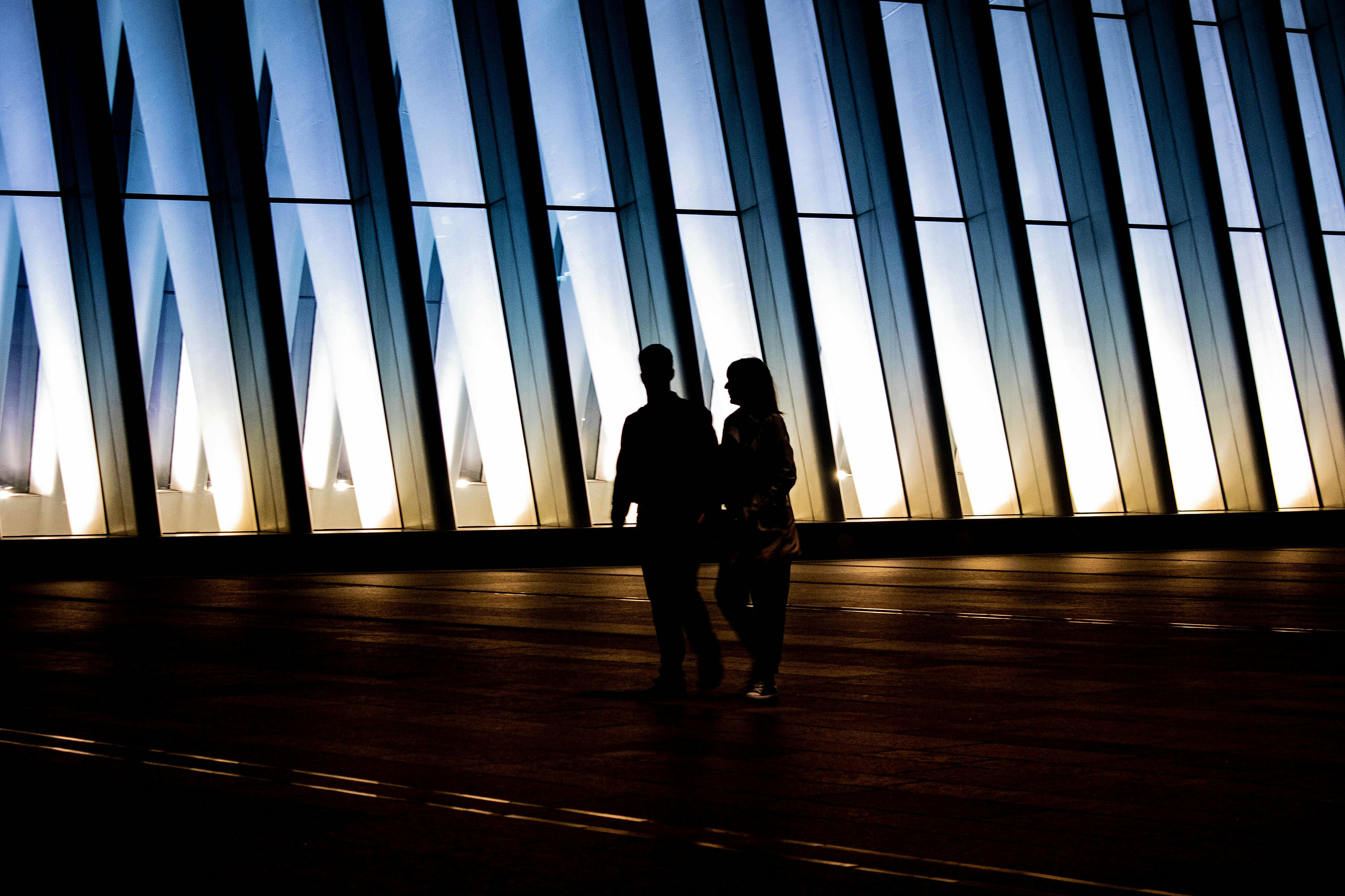Two people walking in front of a modern illuminated glass facade at dusk