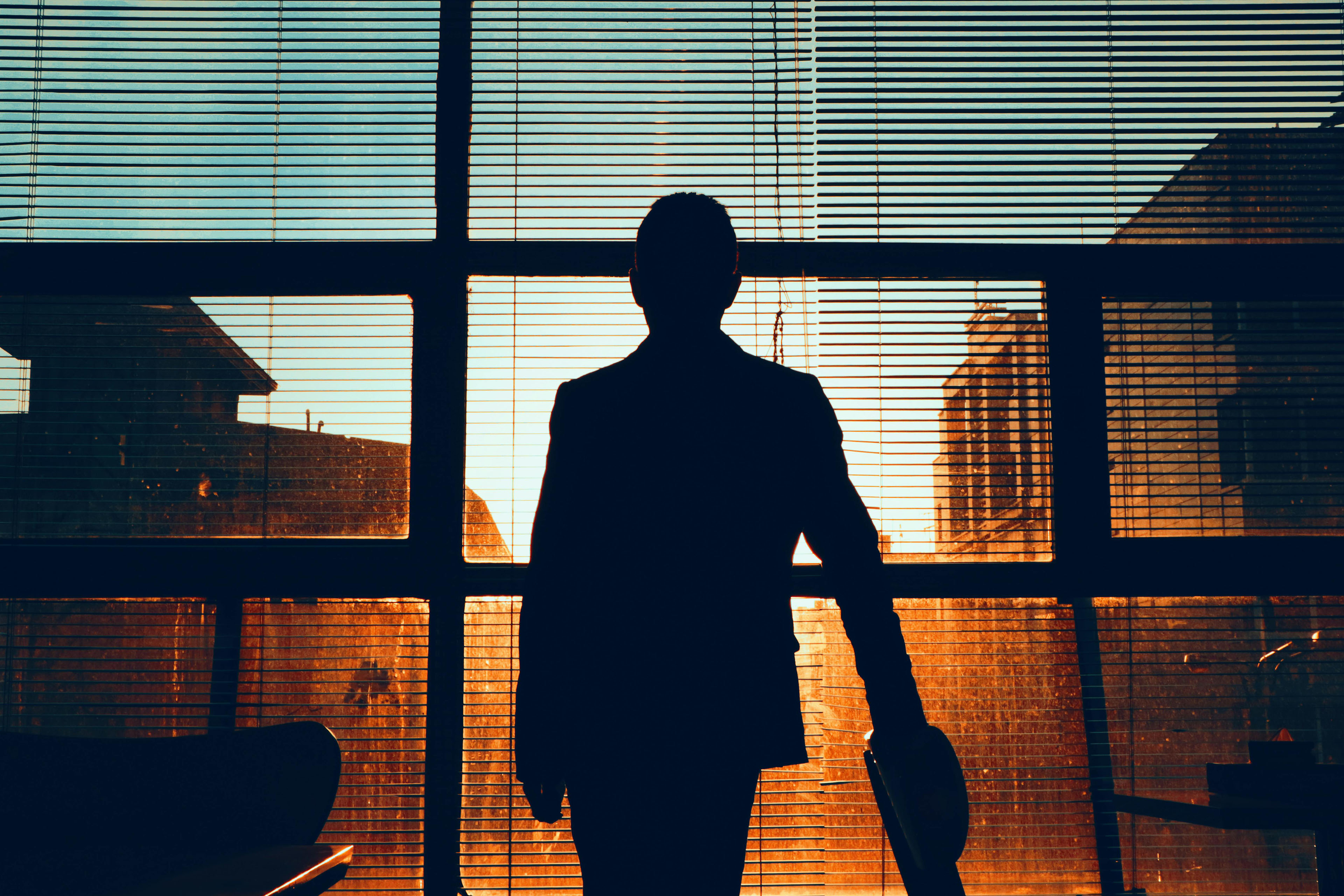 Silhouette of a person standing in front of large windows with blinds, looking at buildings during sunset