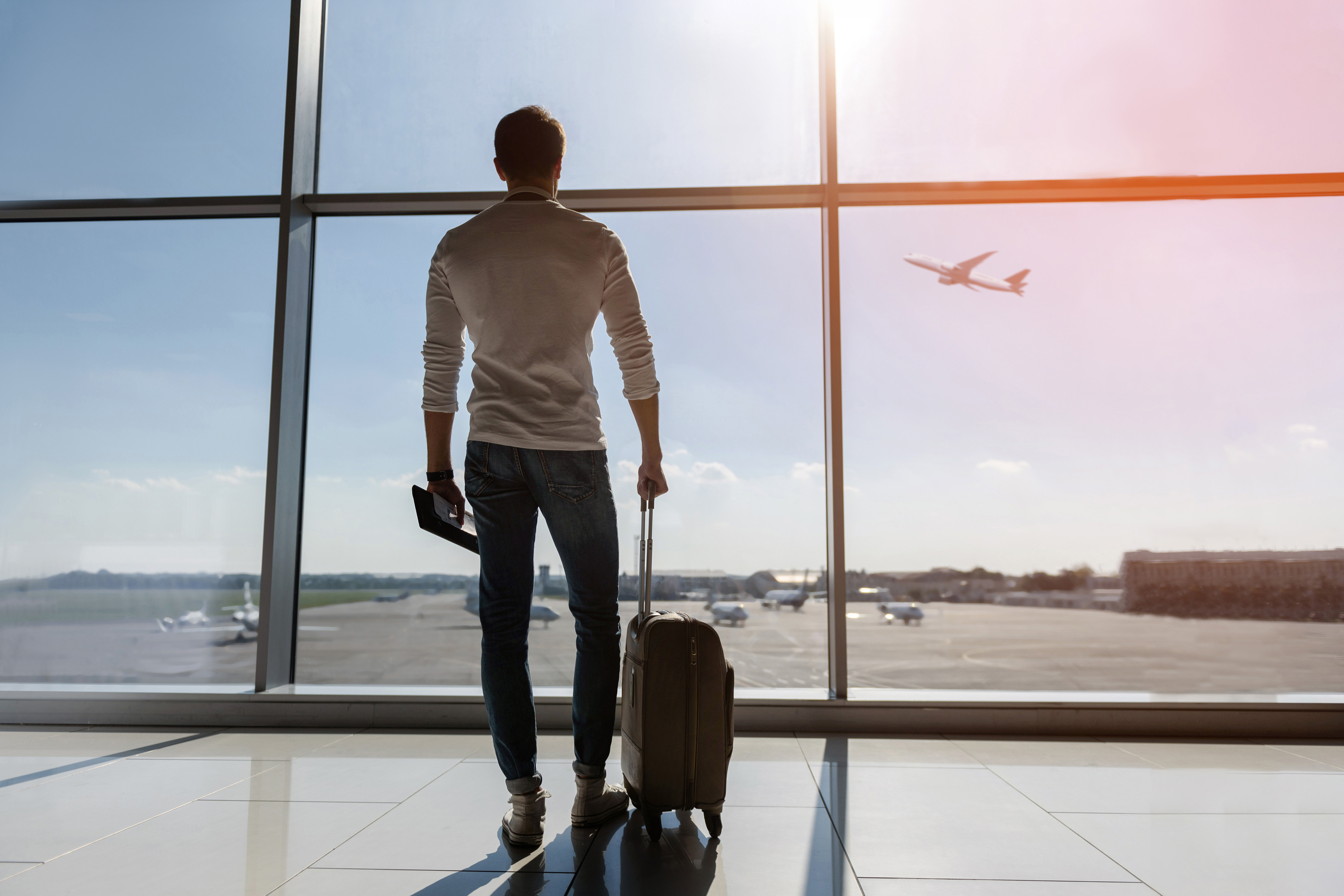 Man stands by an airport window with a suitcase, watching a plane on the runway.