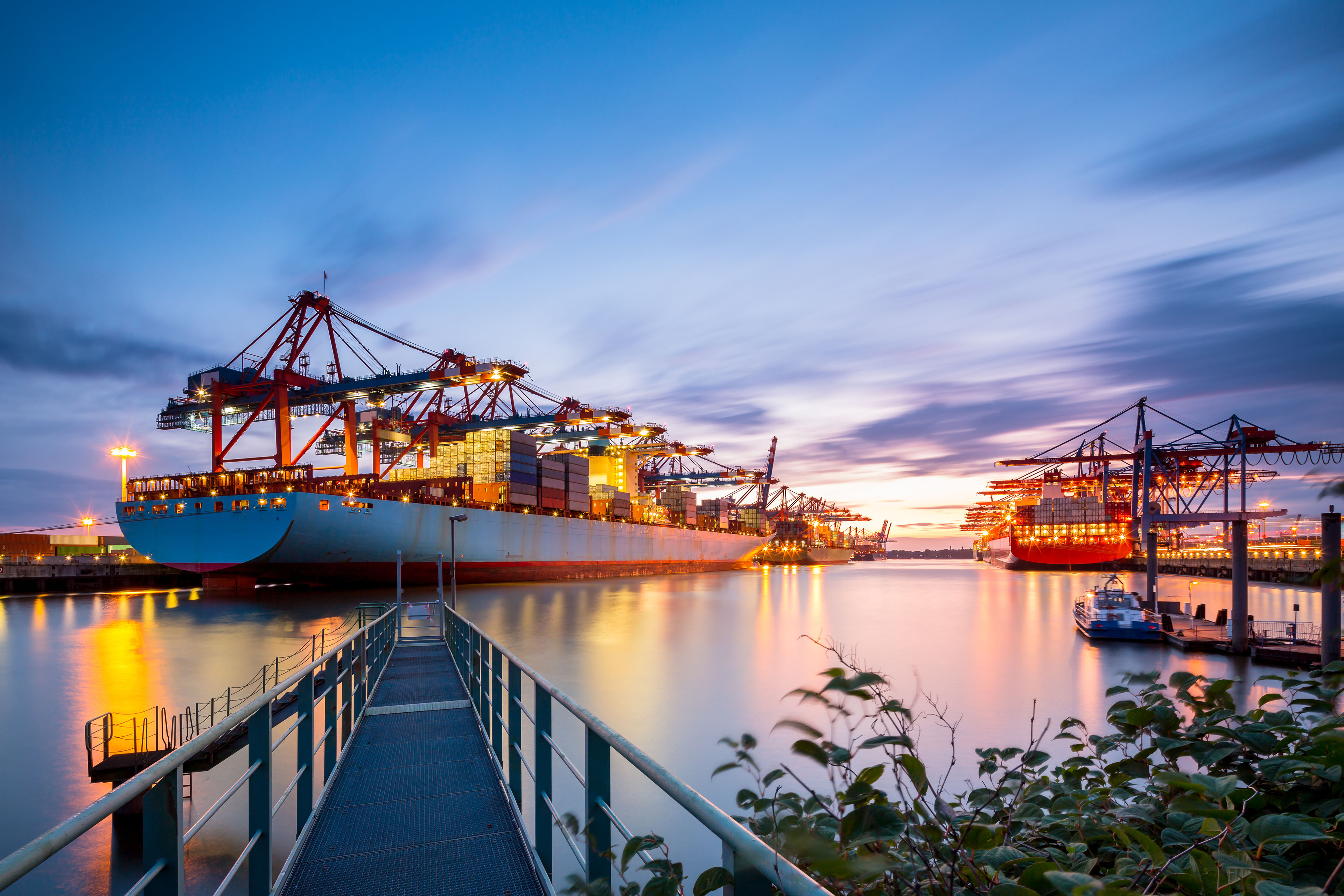 Container ships docked at a port with cranes and lights reflecting on the water at dusk.