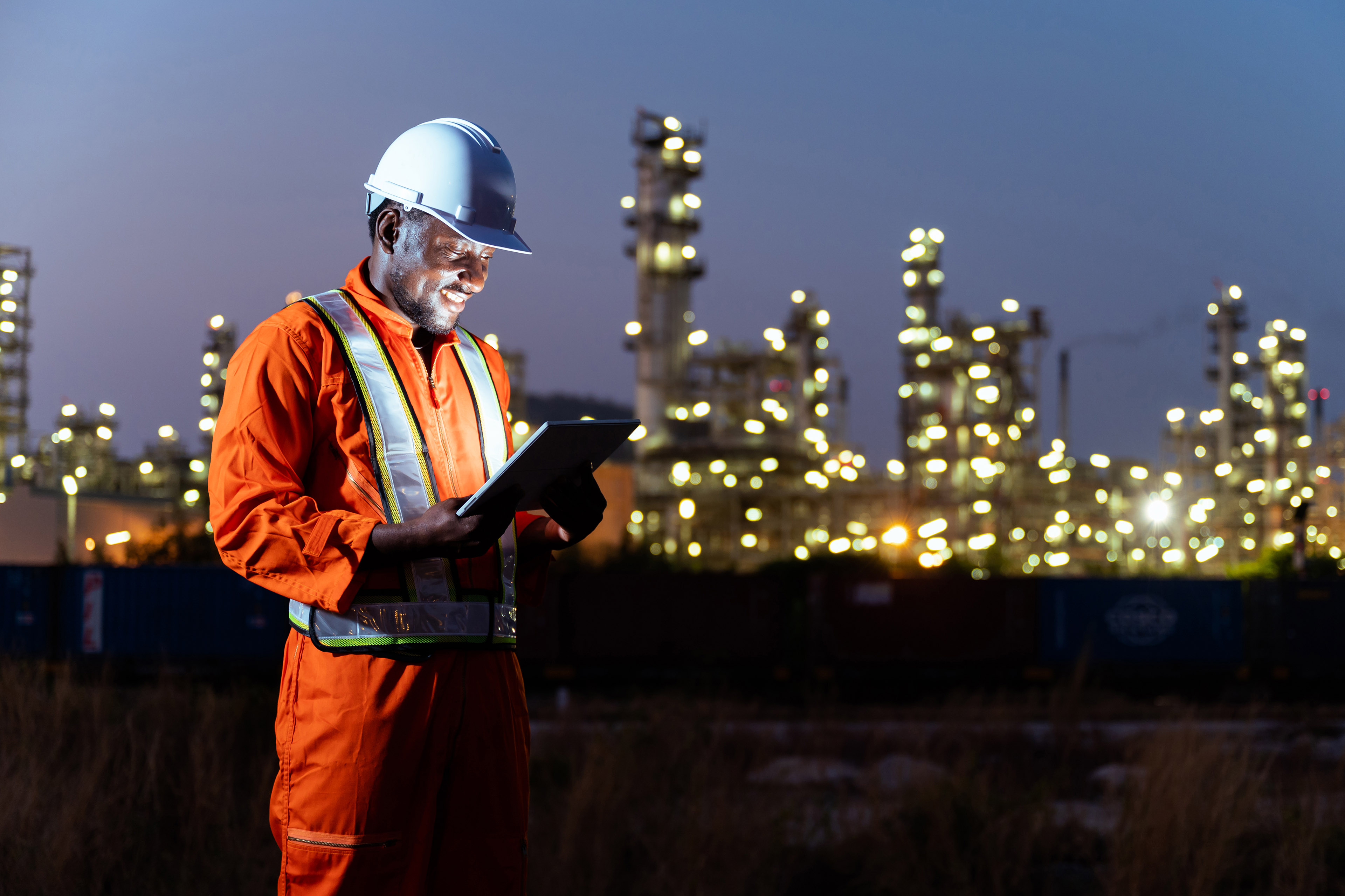 Construction worker using a tablet at an industrial site at night with illuminated facilities in the background