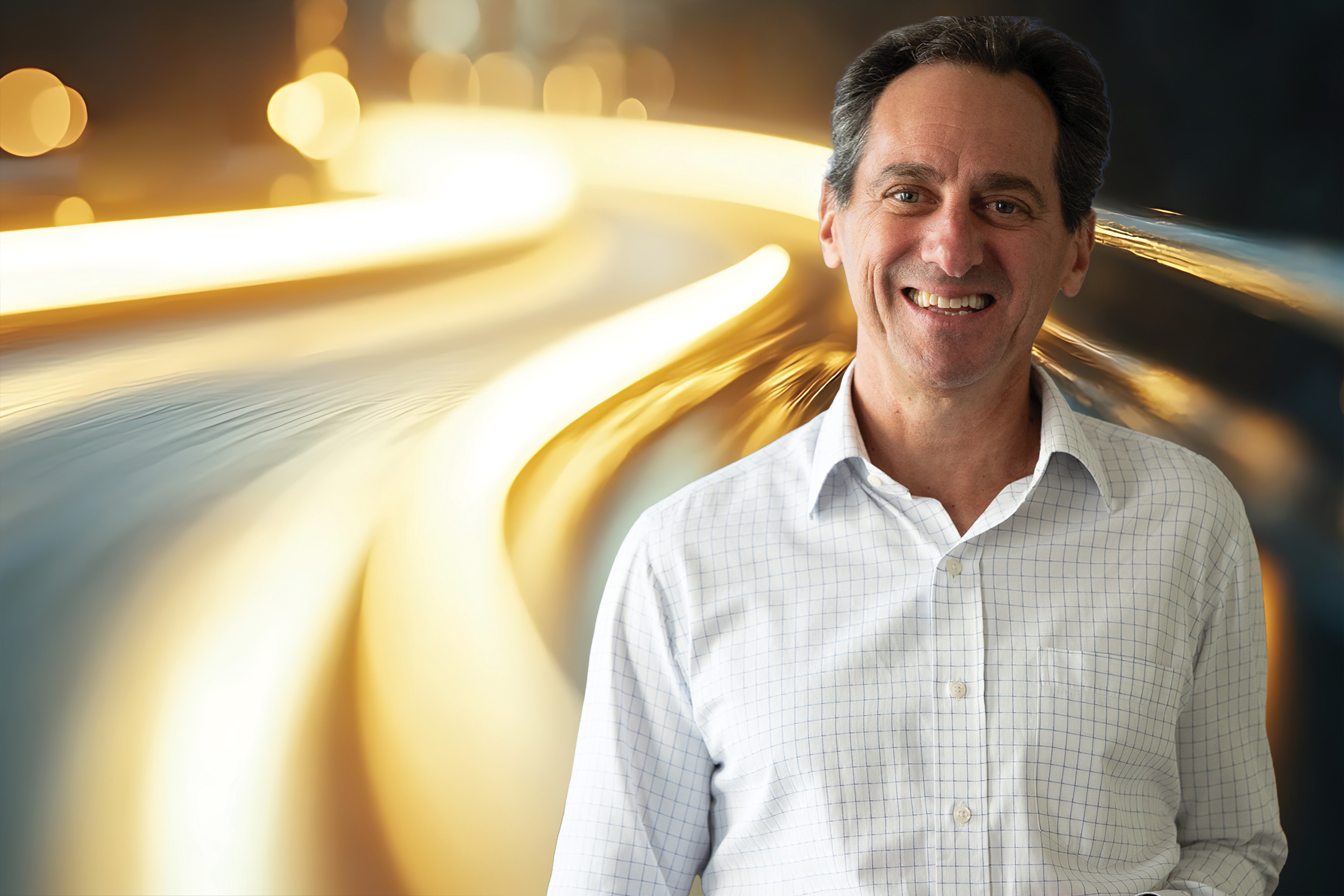 Smiling man in a light-colored shirt standing beside flowing golden light trails on a dark background.