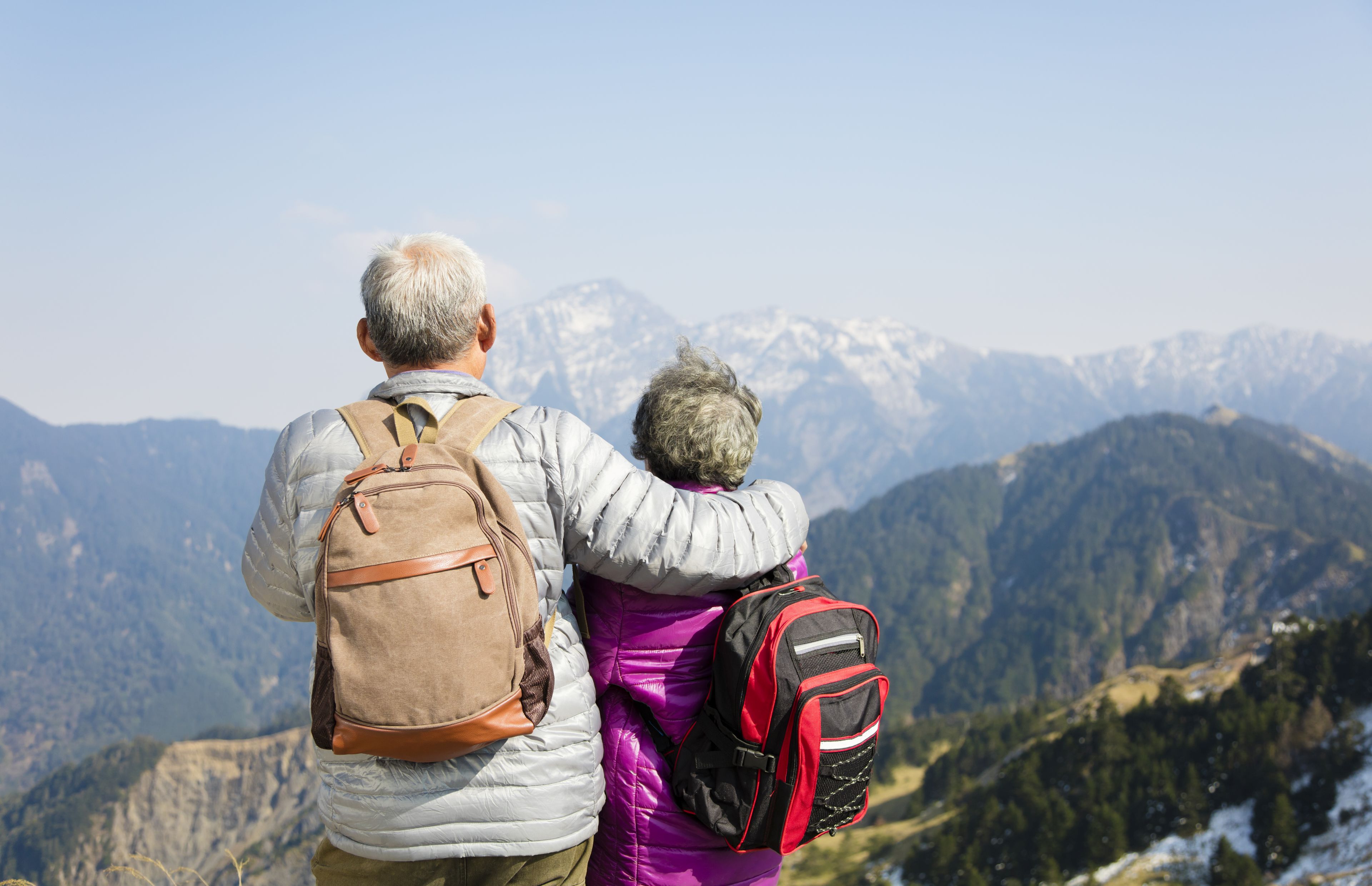Senior couple with backpacks standing on a mountain ridge, overlooking a snowy mountain range