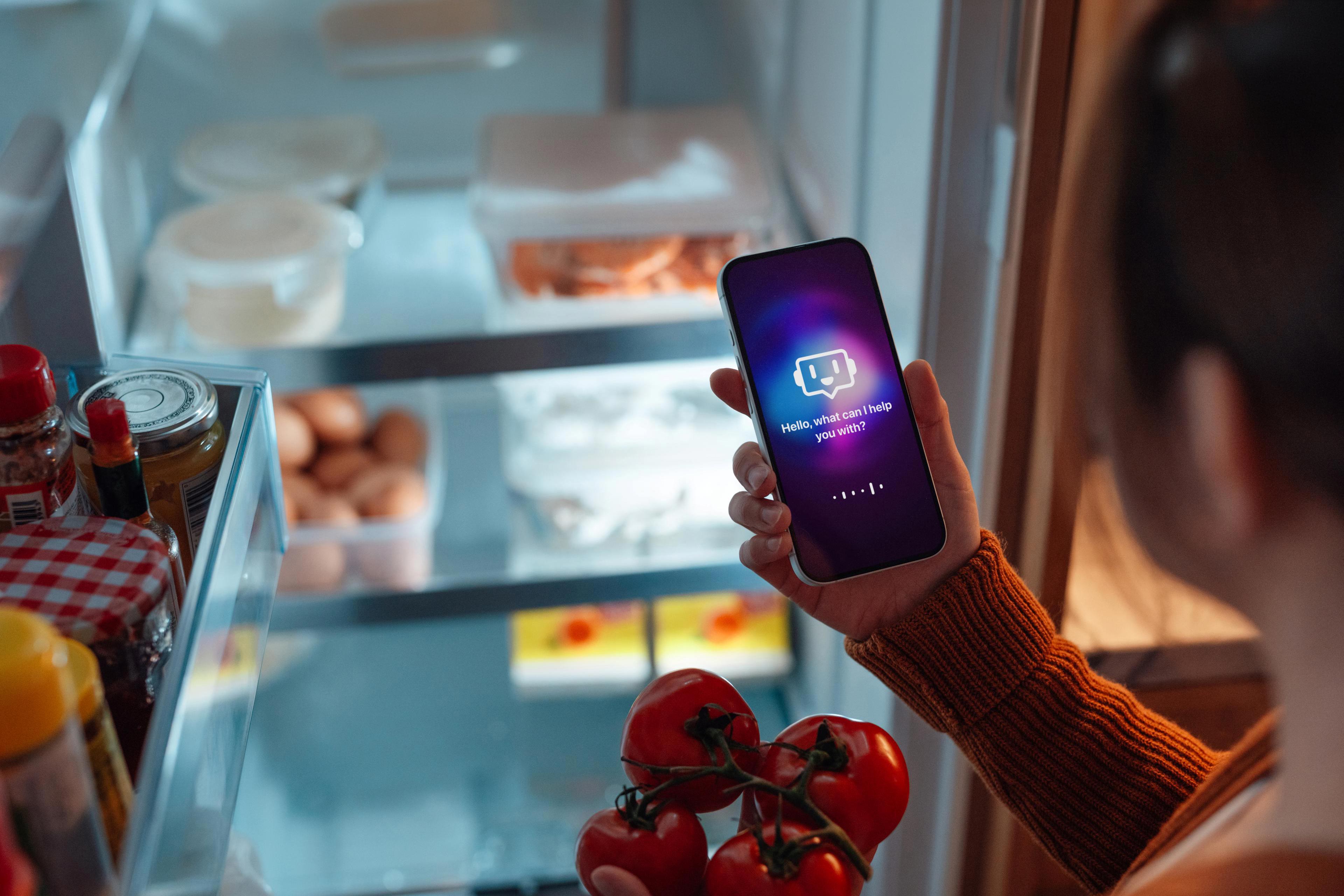 Personne tenant un téléphone intelligent avec une application d’assistant virtuel devant un réfrigérateur ouvert