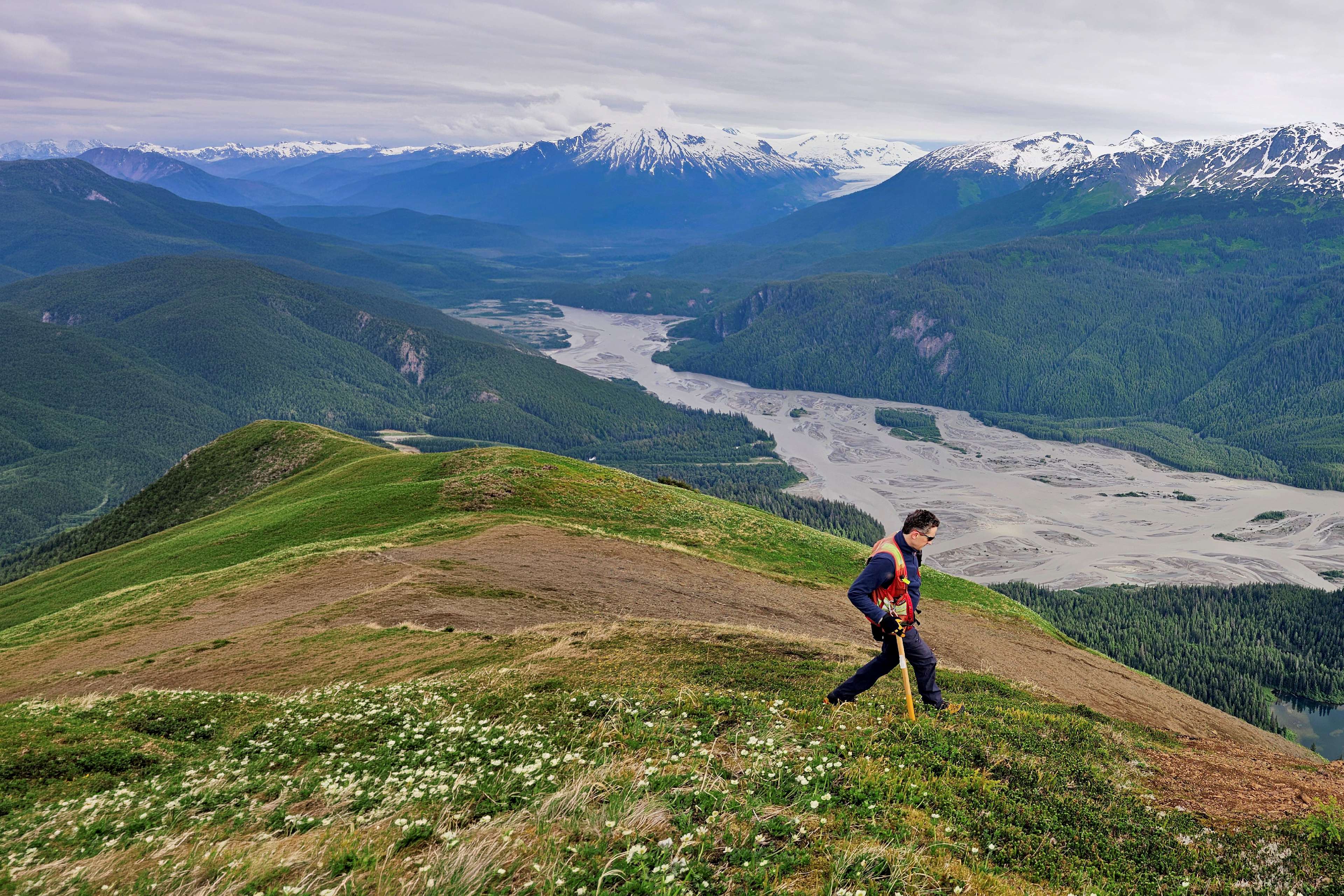 Person hiking on a mountain ridge overlooking a wide valley and snow‑covered peaks