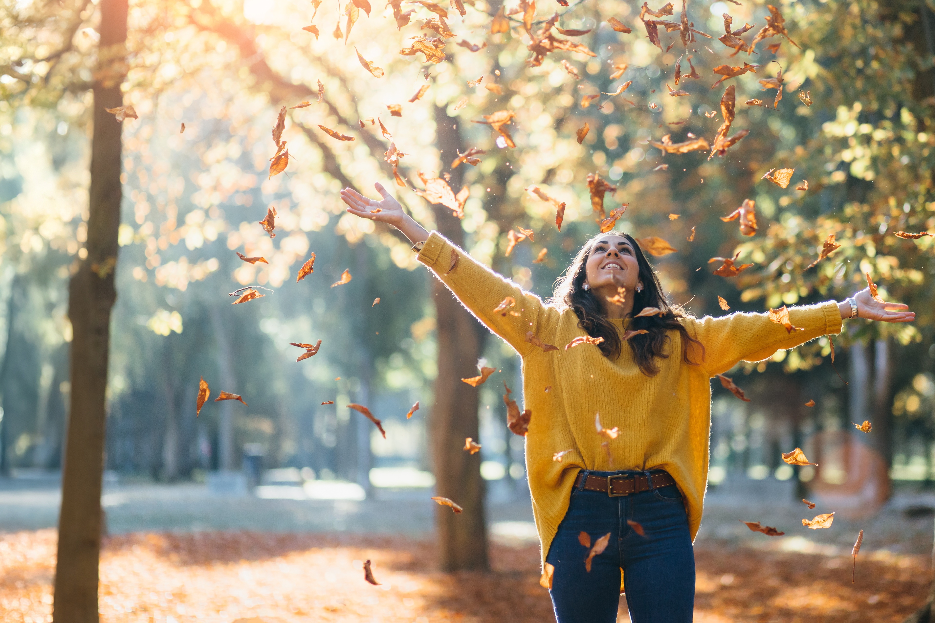 Person in a yellow sweater tossing autumn leaves beneath sunlit trees in a park