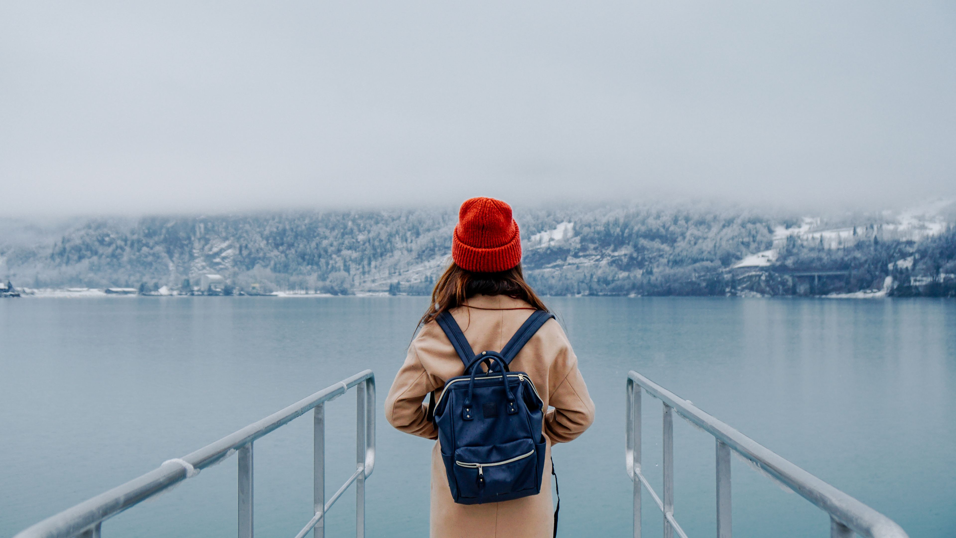 A person wearing a tan coat from behind, carrying a navy blue Anello-Style backpack with a wide opening and top handles