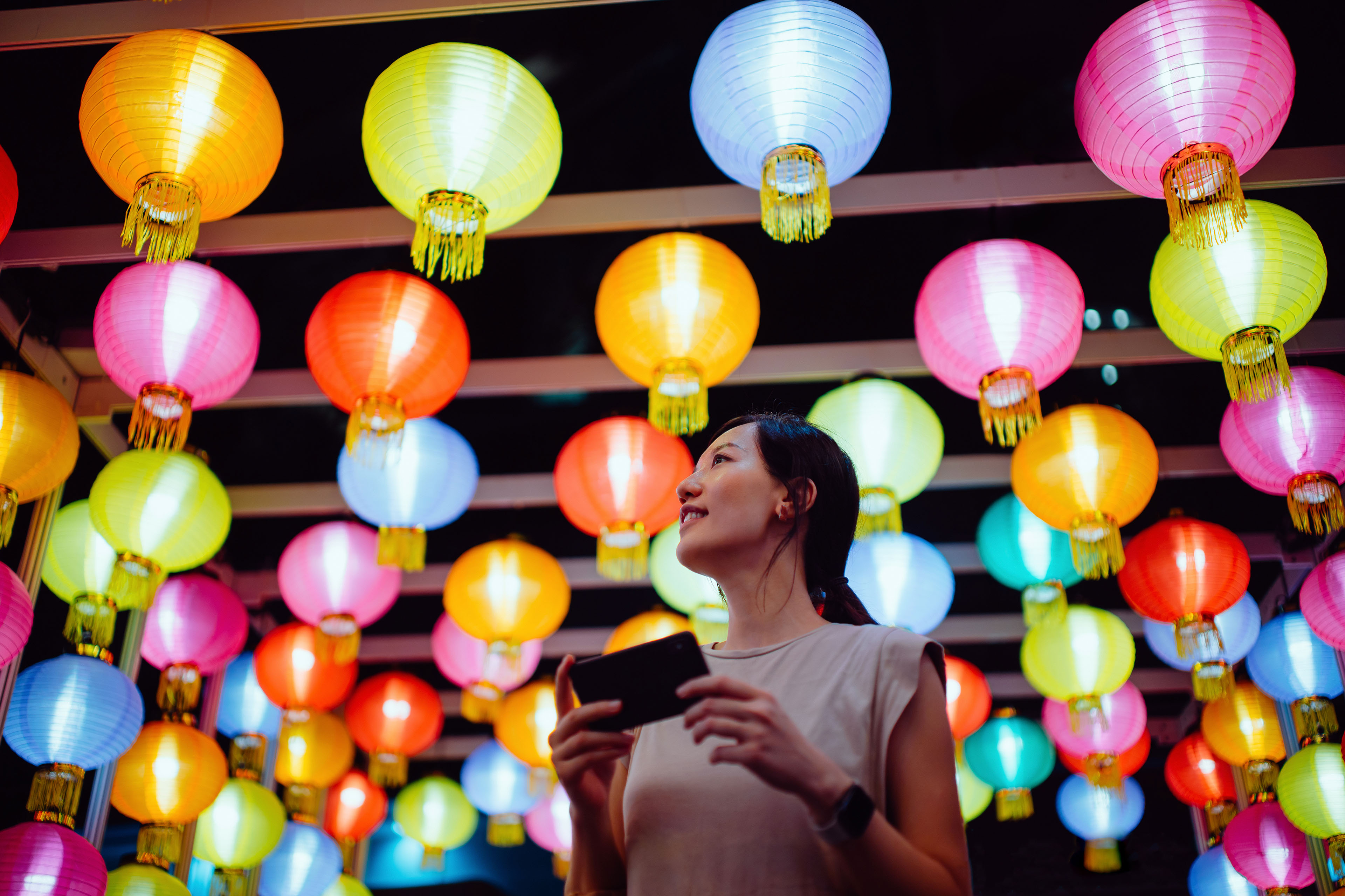 Smiling young Asian woman taking photos of illuminated and colorful traditional Chinese lanterns