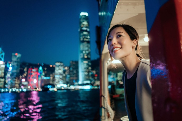 Beautiful smiling young Asian woman looking out through the window, enjoying spectacular illuminated night view 