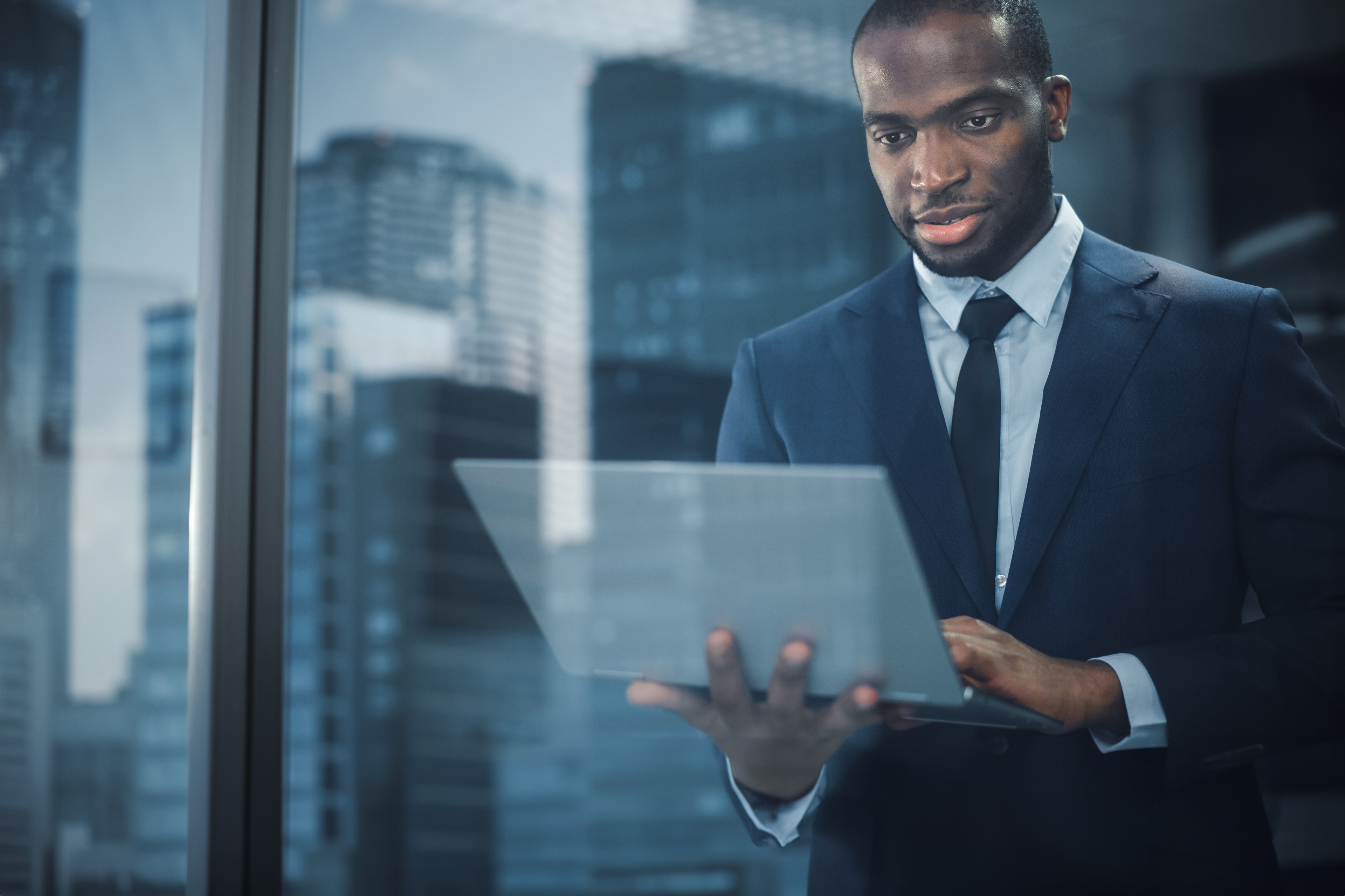 Portrait of Successful Black Businessman Wearing Suit Standing, Using Laptop Looking out of Office Window.