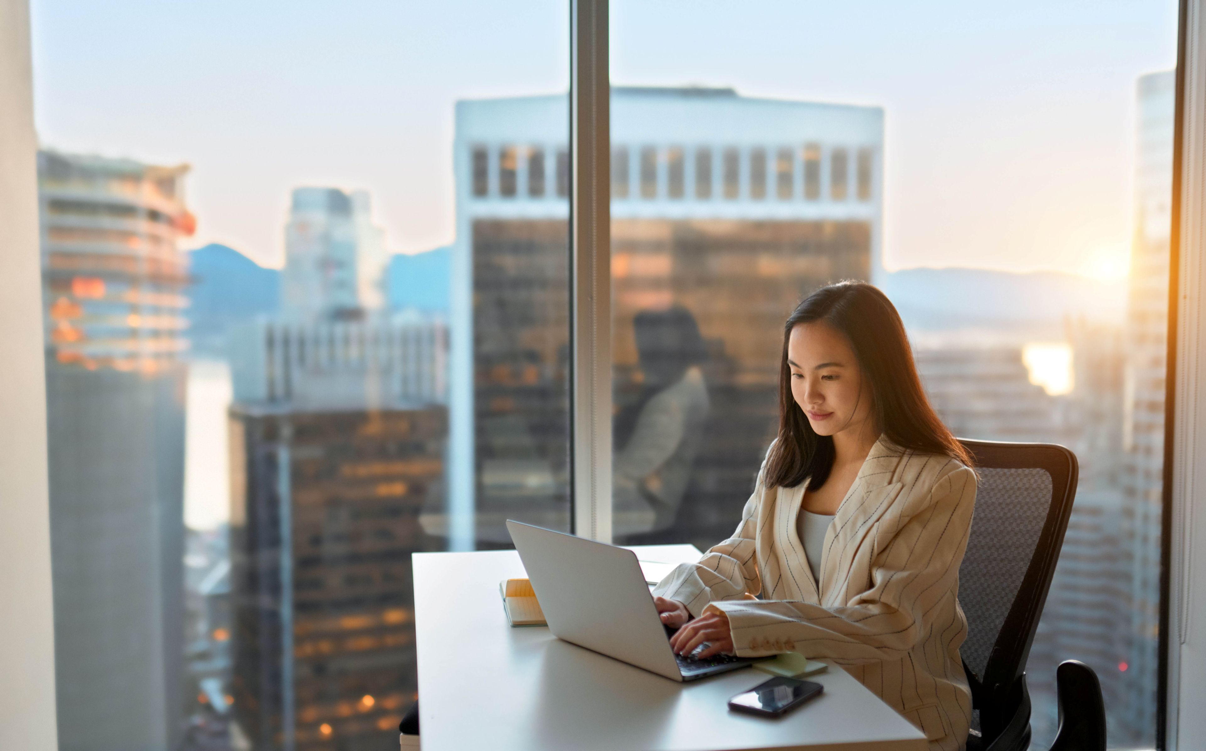 Woman working on a laptop at a desk in a high‑rise office with a city skyline in the background.