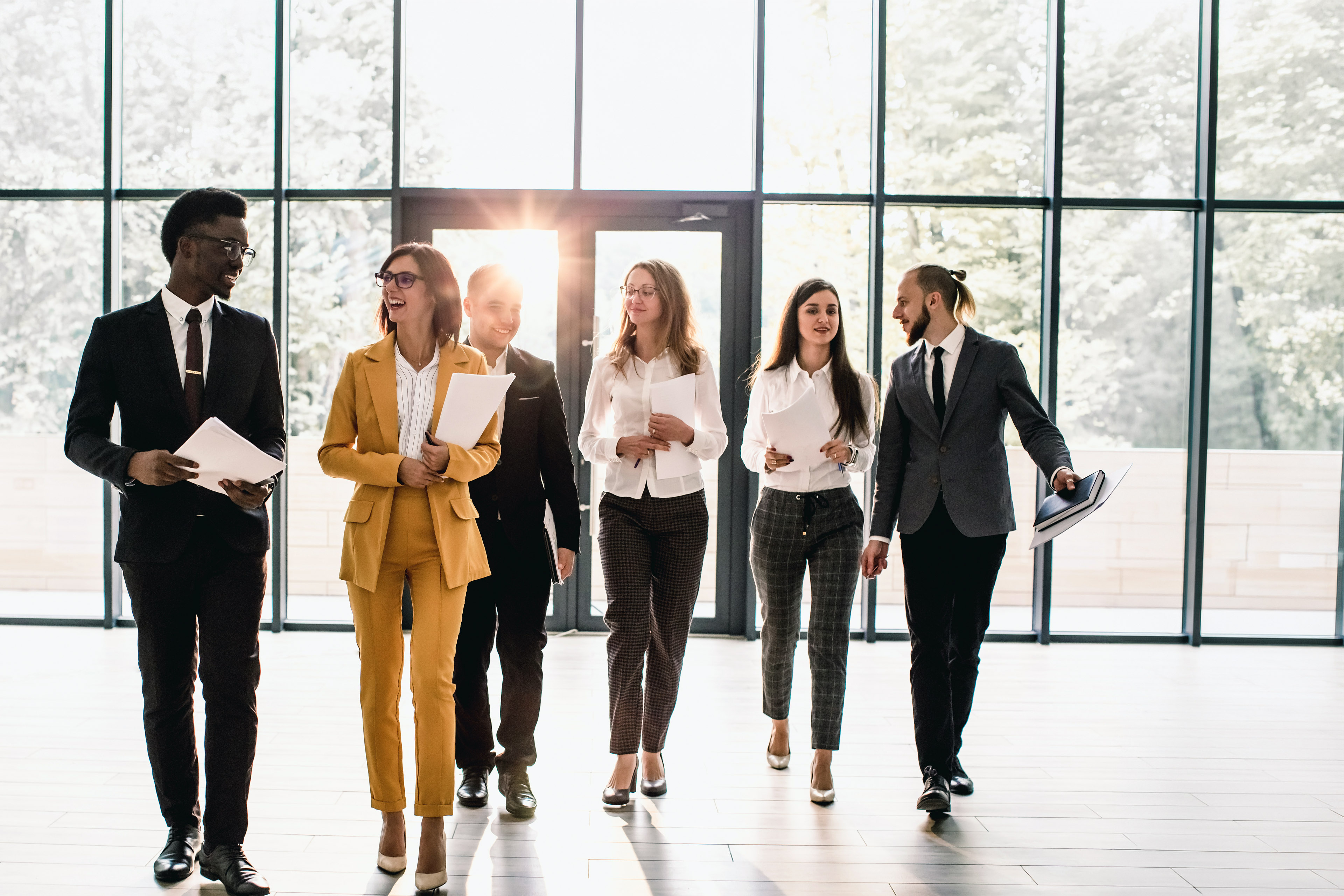 Group of young professionals walking toward camera during a meeting in a modern office with large windows.