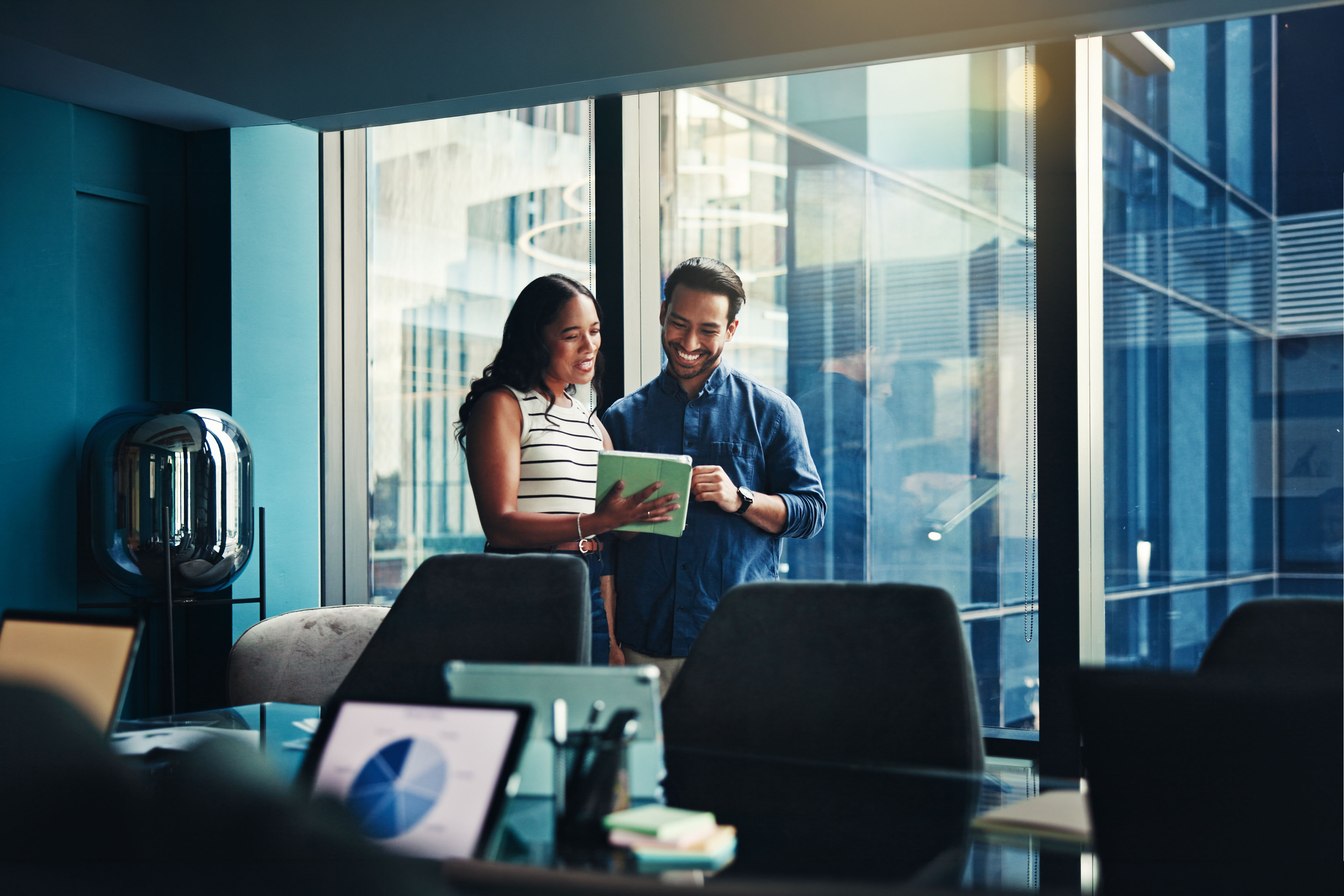 Two professionals reviewing a tablet in a modern conference room with large windows and city views.